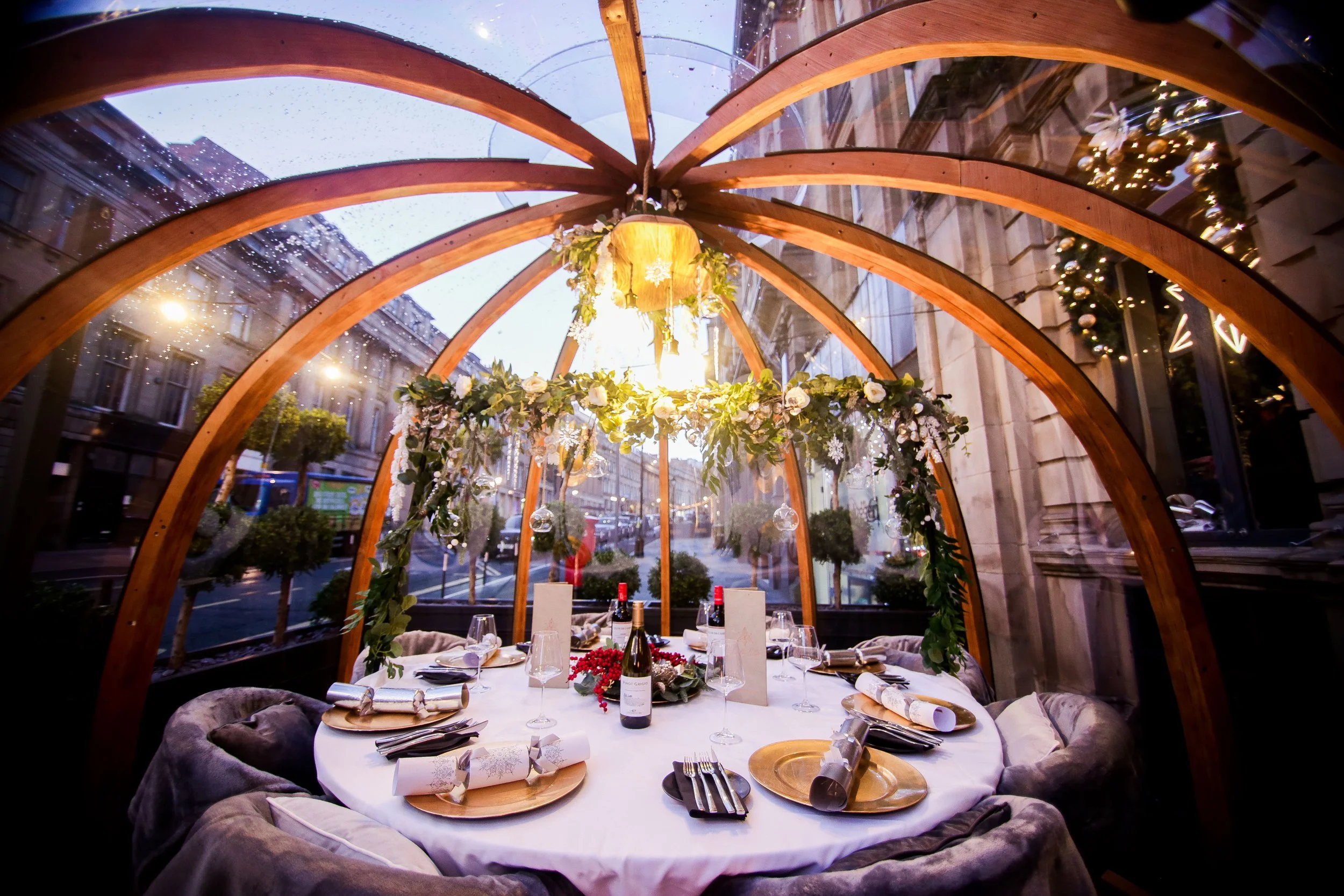 A round table set for a formal dinner, decorated with white tablecloth, gold plates, wine bottles, glasses, and wrapped napkins. The table is within a glass, wooden-domed structure decorated with greenery and white flowers, with city buildings visibl
