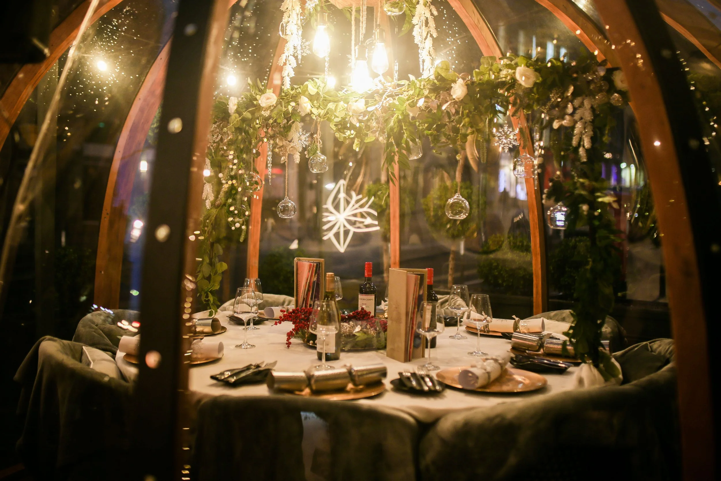 A romantic dinner setup inside a glass-enclosed patio with a round table, decorated with flowers, wine bottles, and wine glasses, illuminated by hanging lights and a star-shaped decoration.