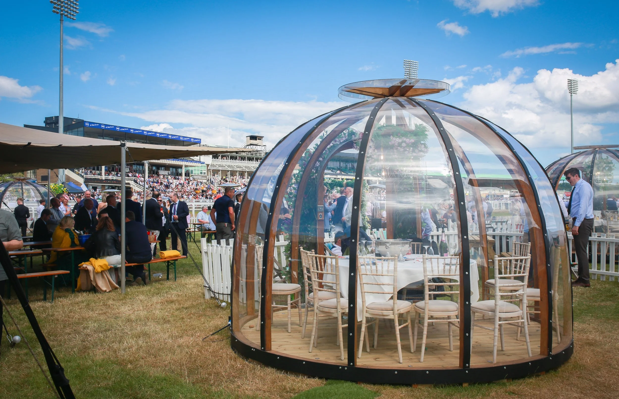 A glass-domed outdoor dining area at a racecourse with a large crowd in the background under a blue sky with clouds.