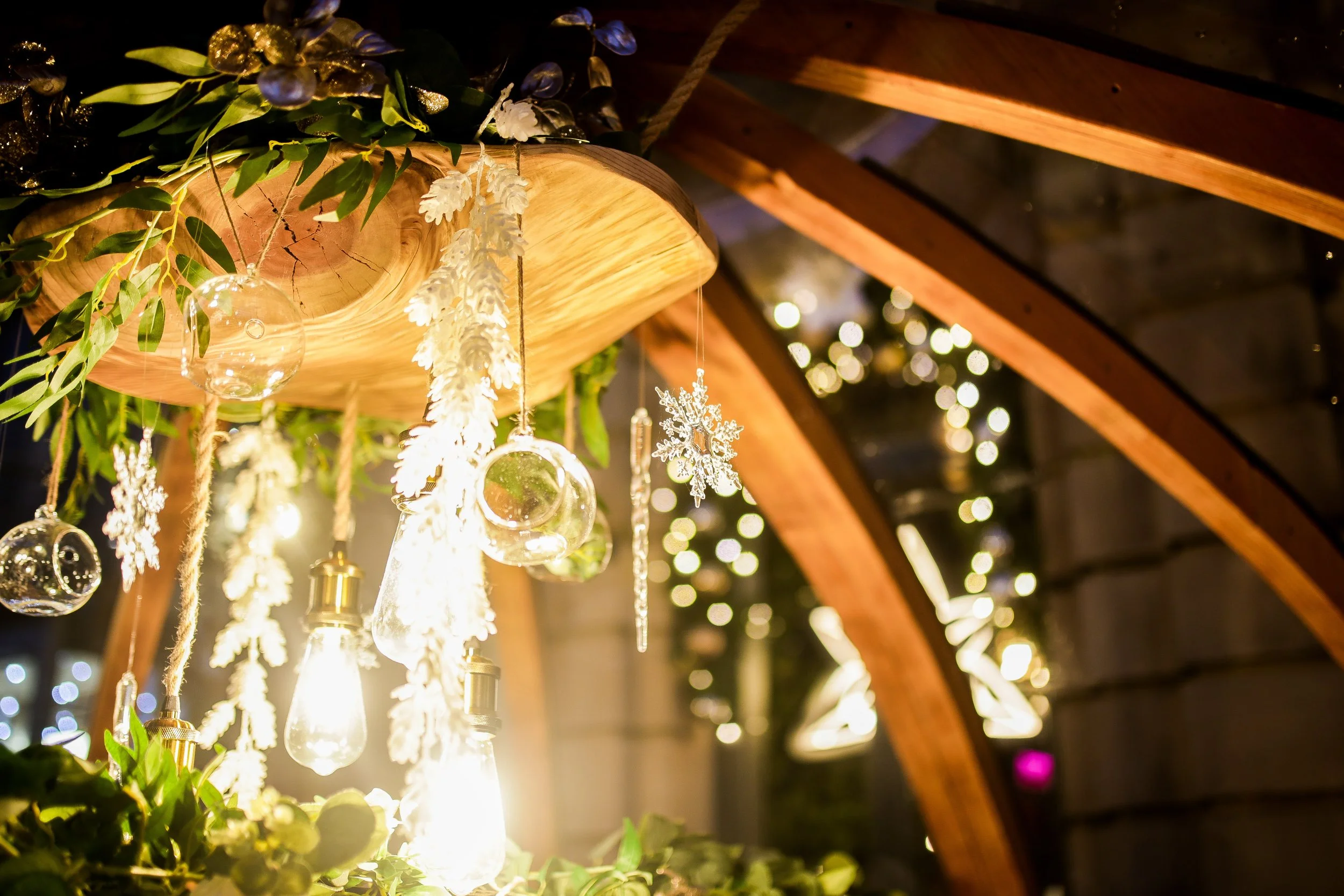Decorative Christmas display with hanging glass ornaments, snowflake ornaments, and greenery inside a wooden and glass structure at night.
