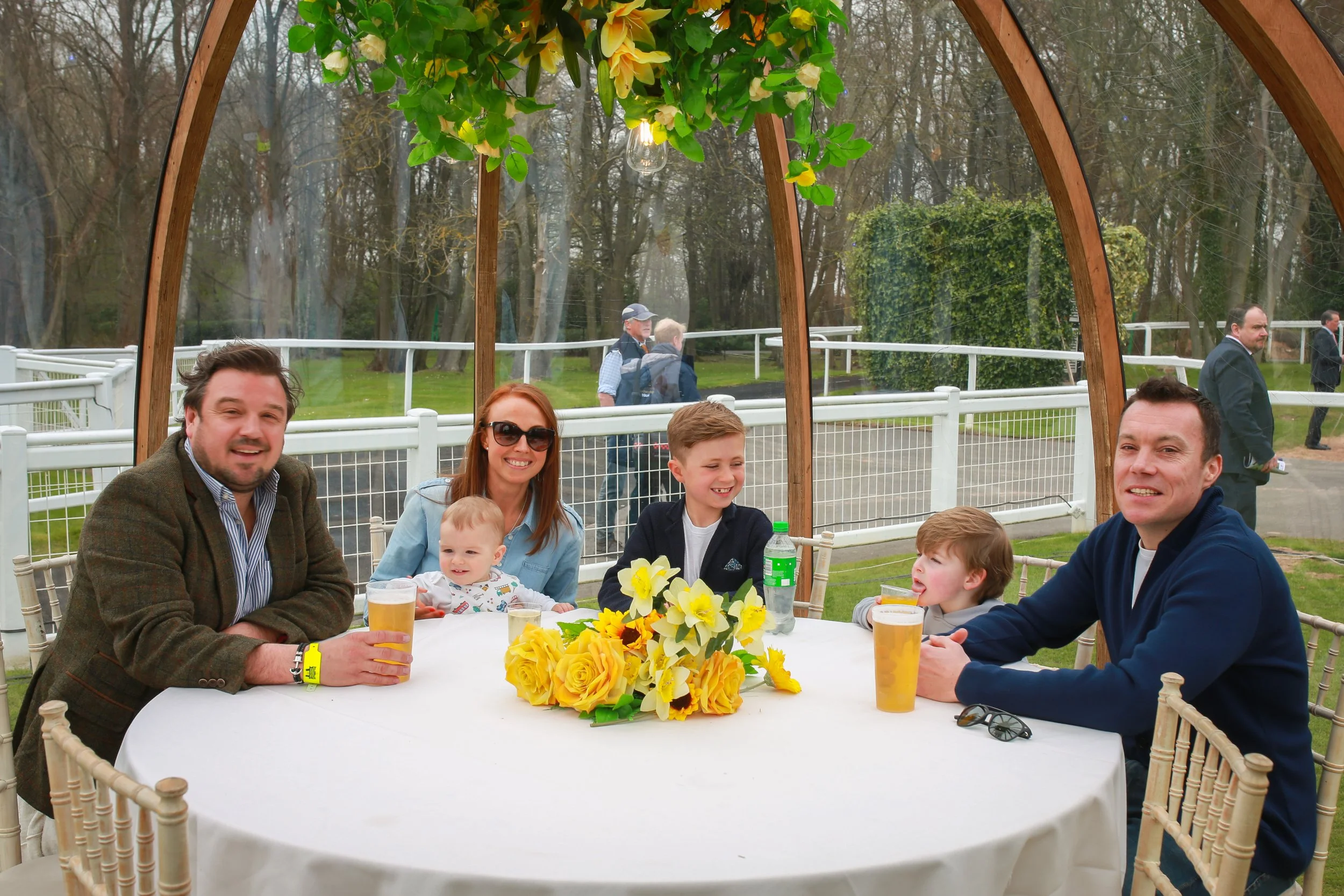 Family enjoying a meal at a round table with a yellow flower centerpiece under a wooden gazebo at outdoor event.