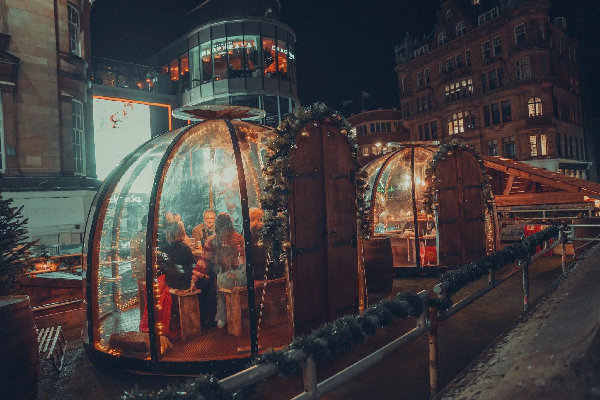 Children inside glass igloos decorated for Christmas, sitting at tables, at night with festive lights and holiday decorations.