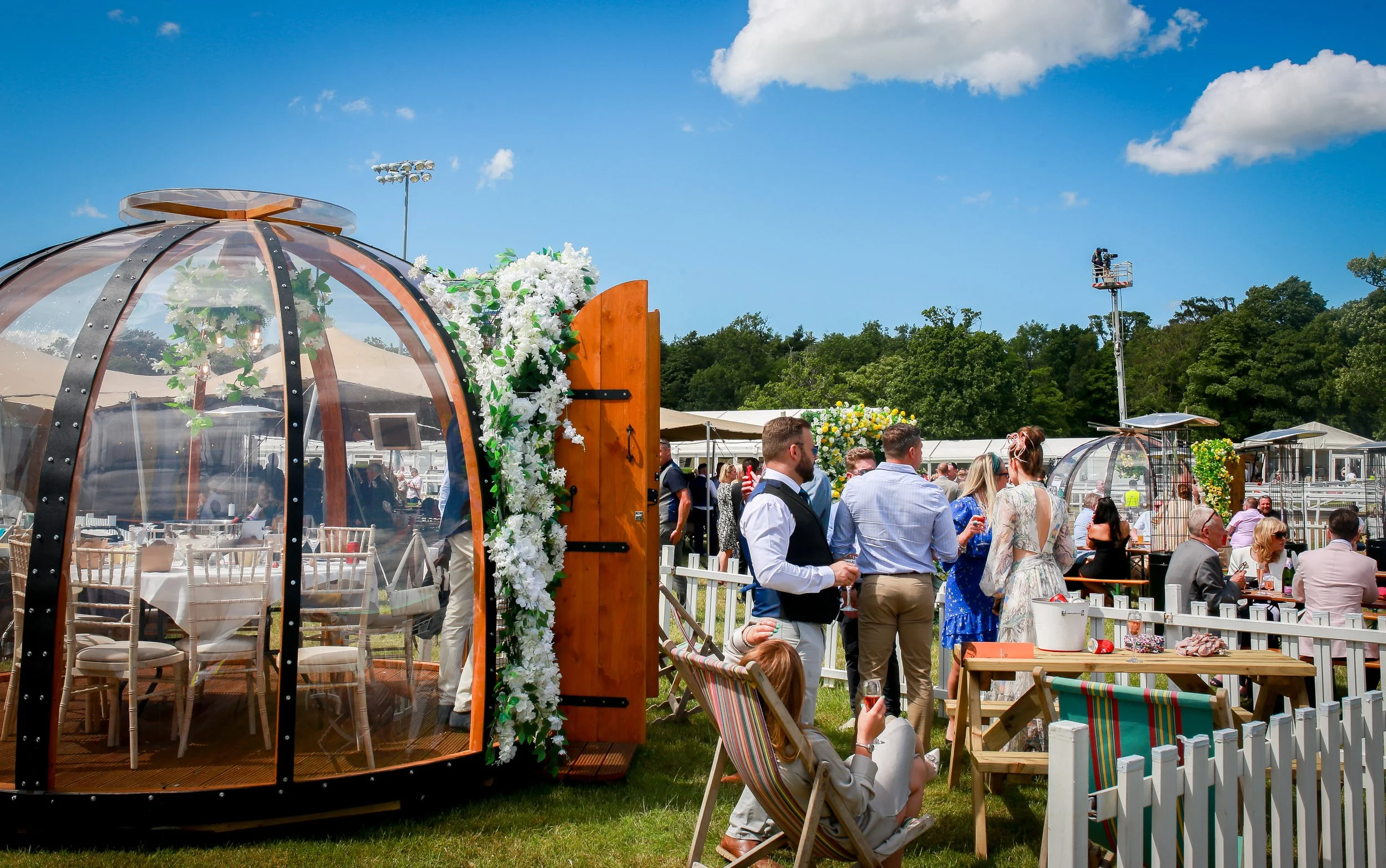 People gather at an outdoor wedding reception, with a transparent, dome-shaped wine tasting tent decorated with flowers in the foreground, and trees and a clear blue sky in the background.