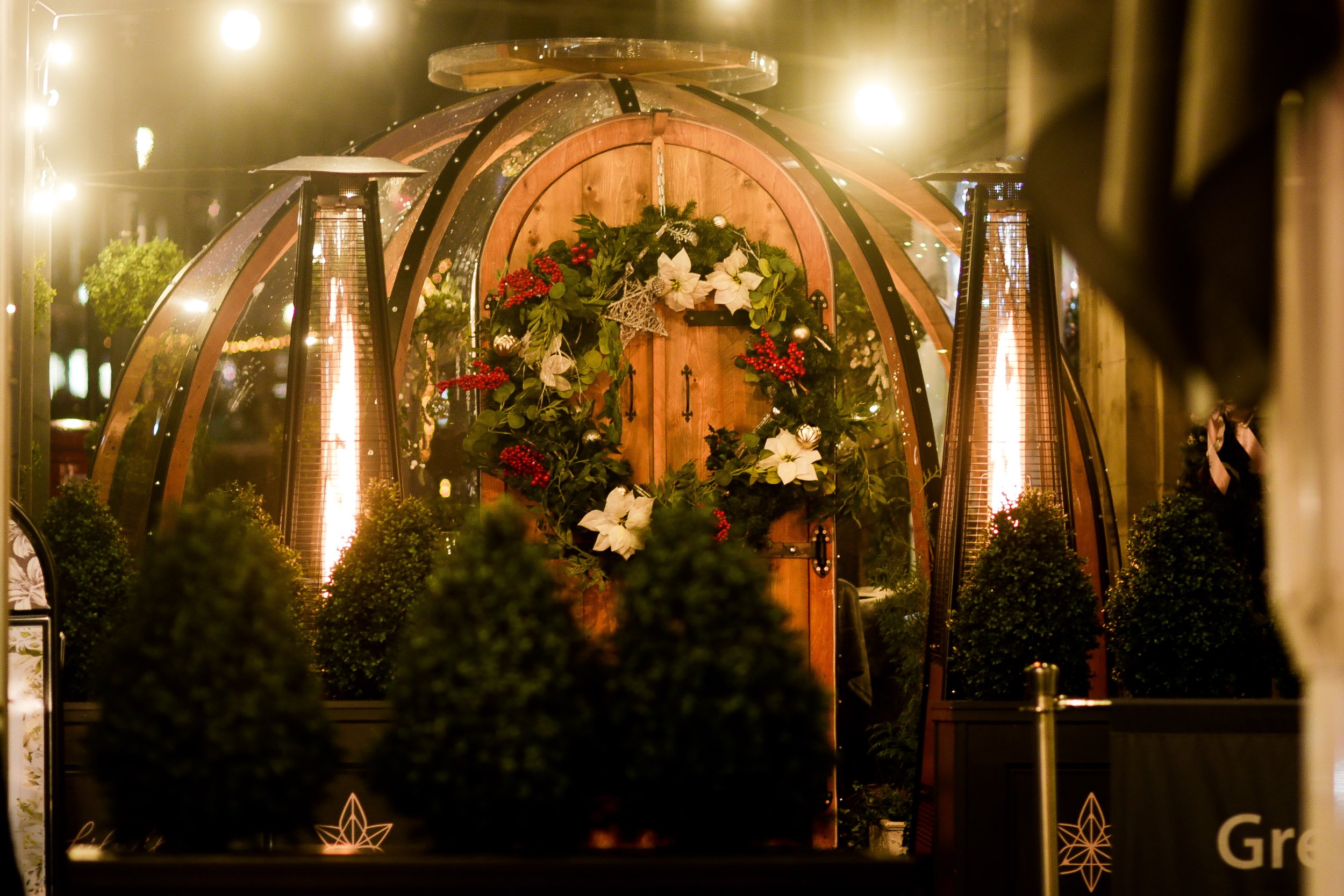 A Christmas wreath decorated with white poinsettias, red berries, and greenery hanging on a rustic wooden door inside a glass-domed structure, illuminated by warm lighting.