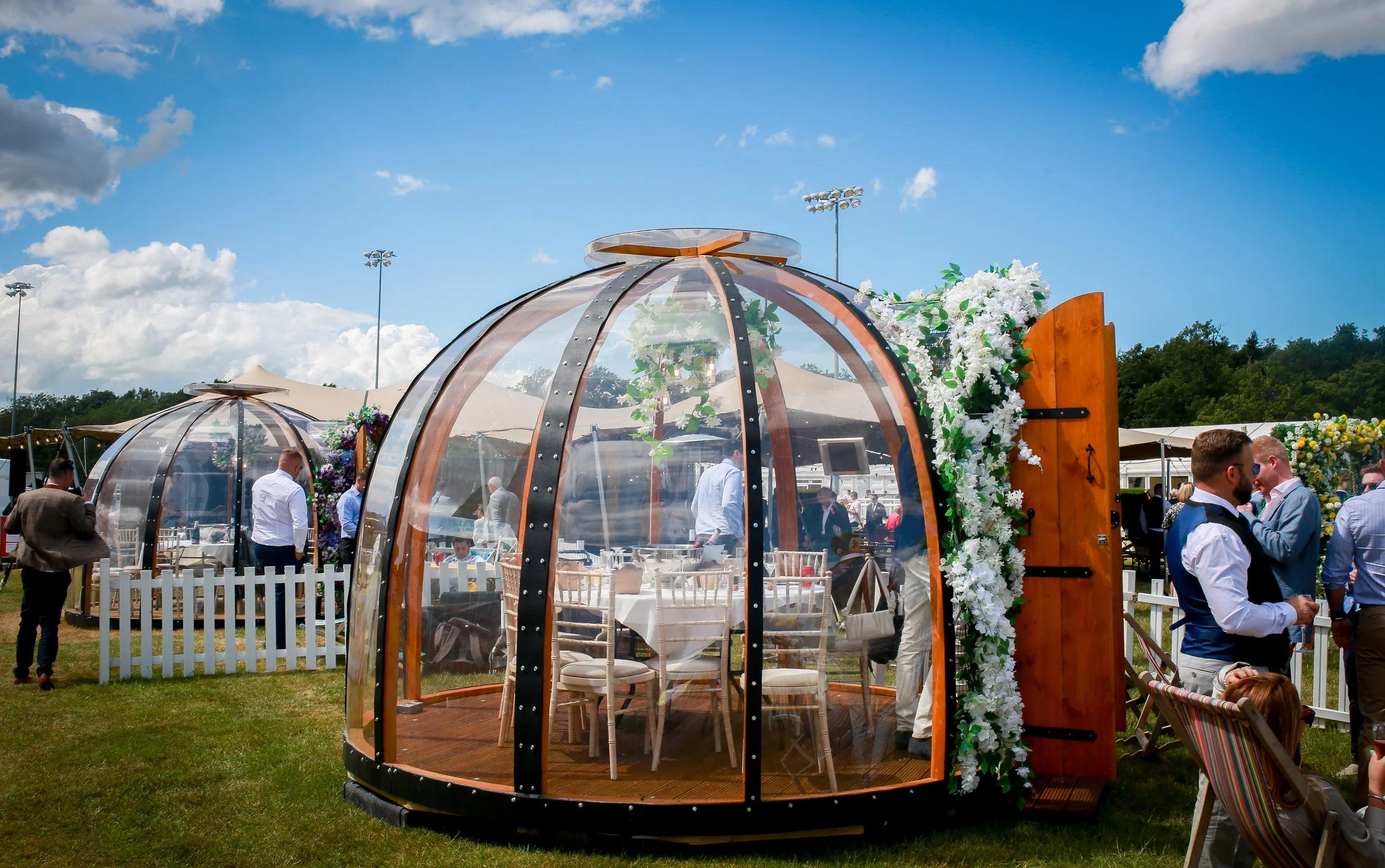 Outdoor wedding reception with glass bubble-shaped tents, decorated with flowers, under a blue sky with clouds, and guests dressed in formal attire.