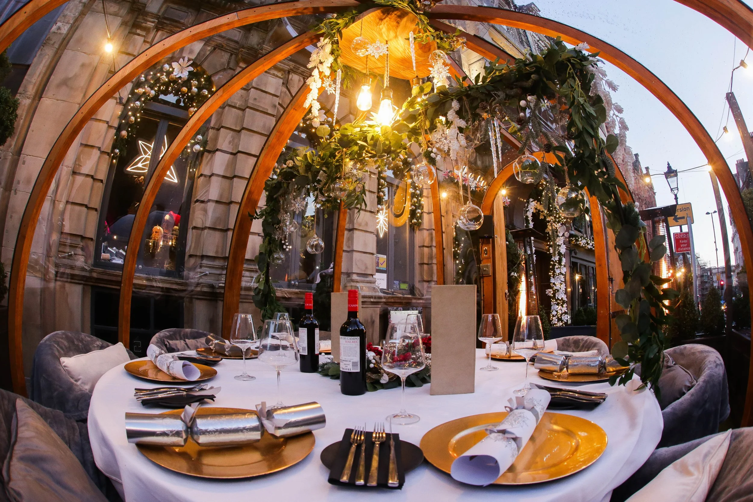 Decorated indoor dining table set for a special occasion with gold plates, wine glasses, silverware, and Christmas-themed decorations, including garlands, hanging ornaments, and string lights, inside a wooden arched structure with large windows.
