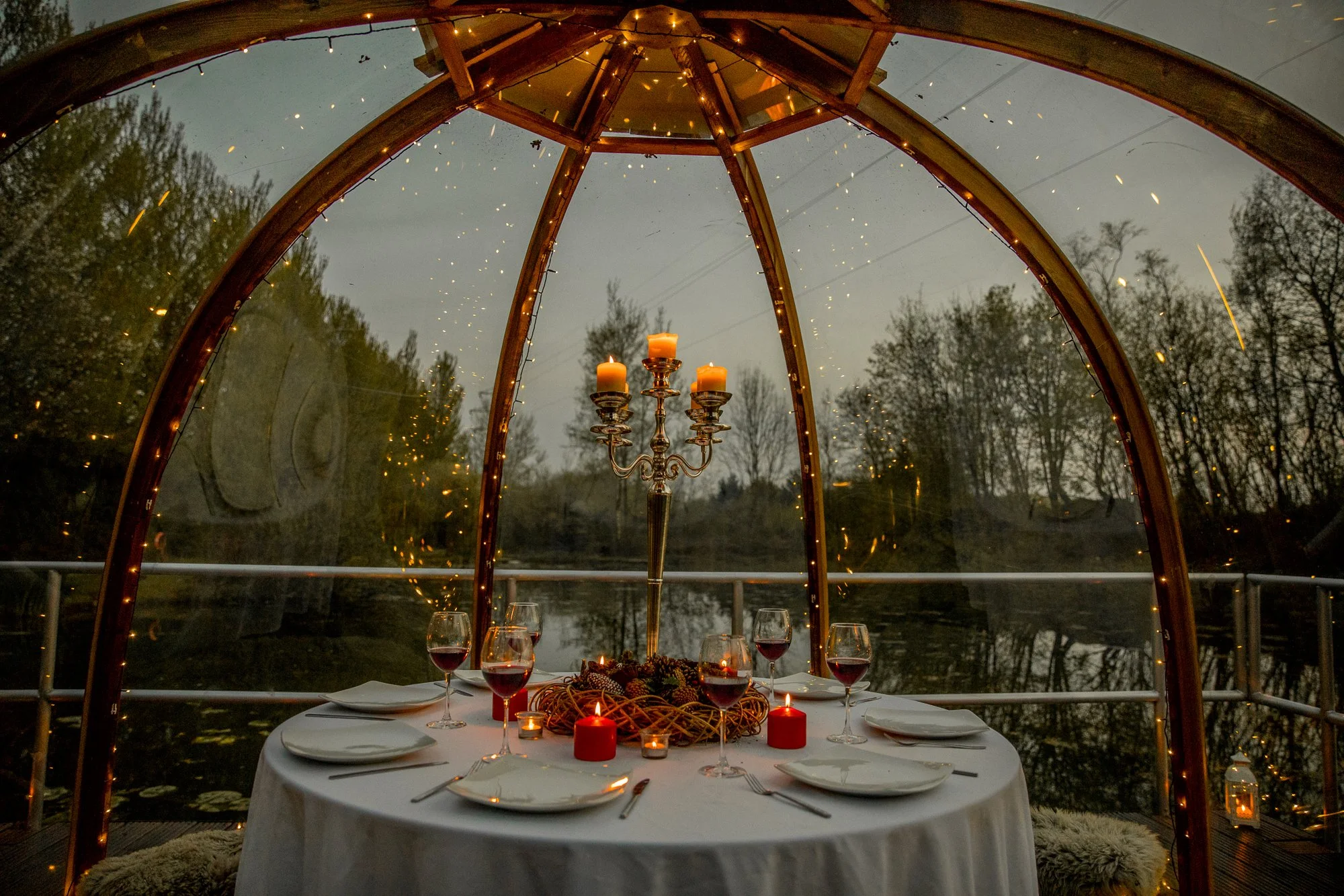 A romantic outdoor dinner setup with a round table covered with a white tablecloth, six wine glasses filled with red wine, and six place settings with white plates and silverware. The table is decorated with a tall candelabrum, several candles, and a