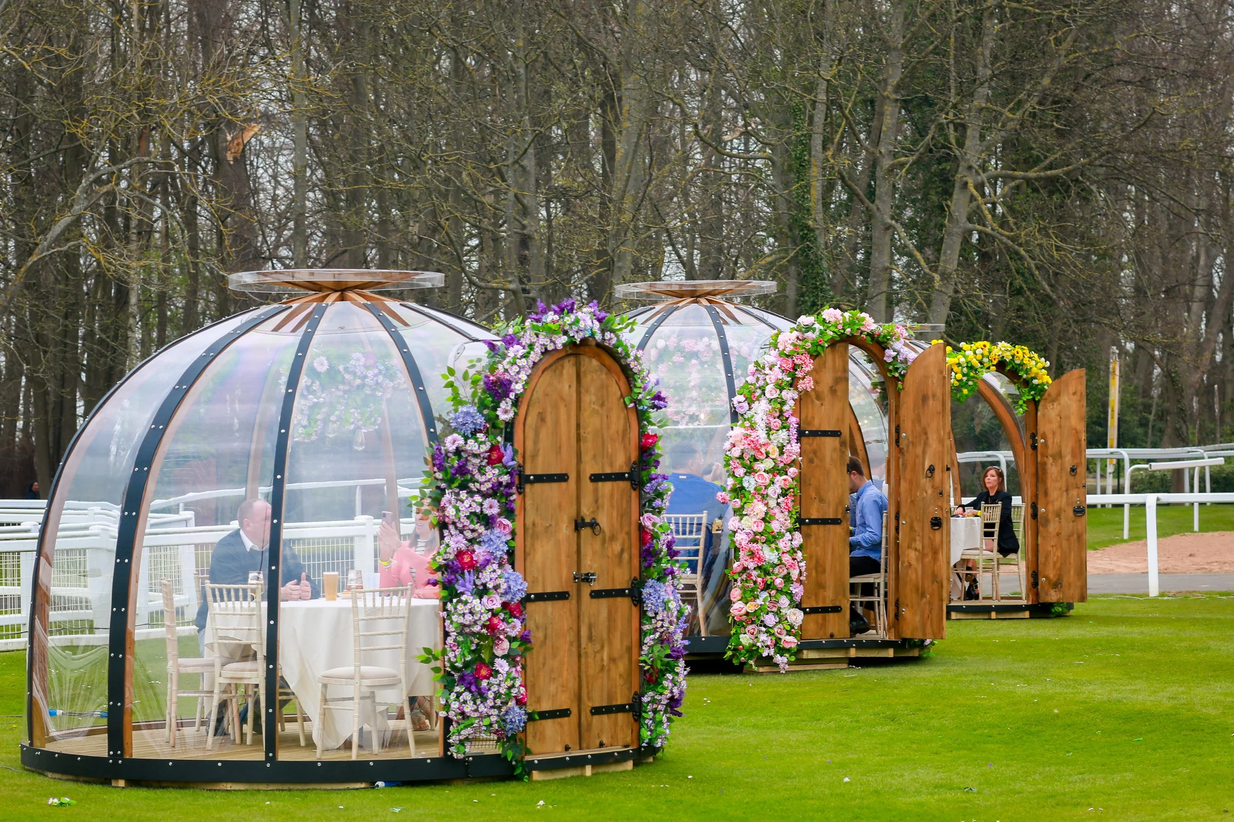Two transparent geodesic dome structures decorated with colorful flowers, serving as outdoor dining areas on a grassy field with trees in the background.