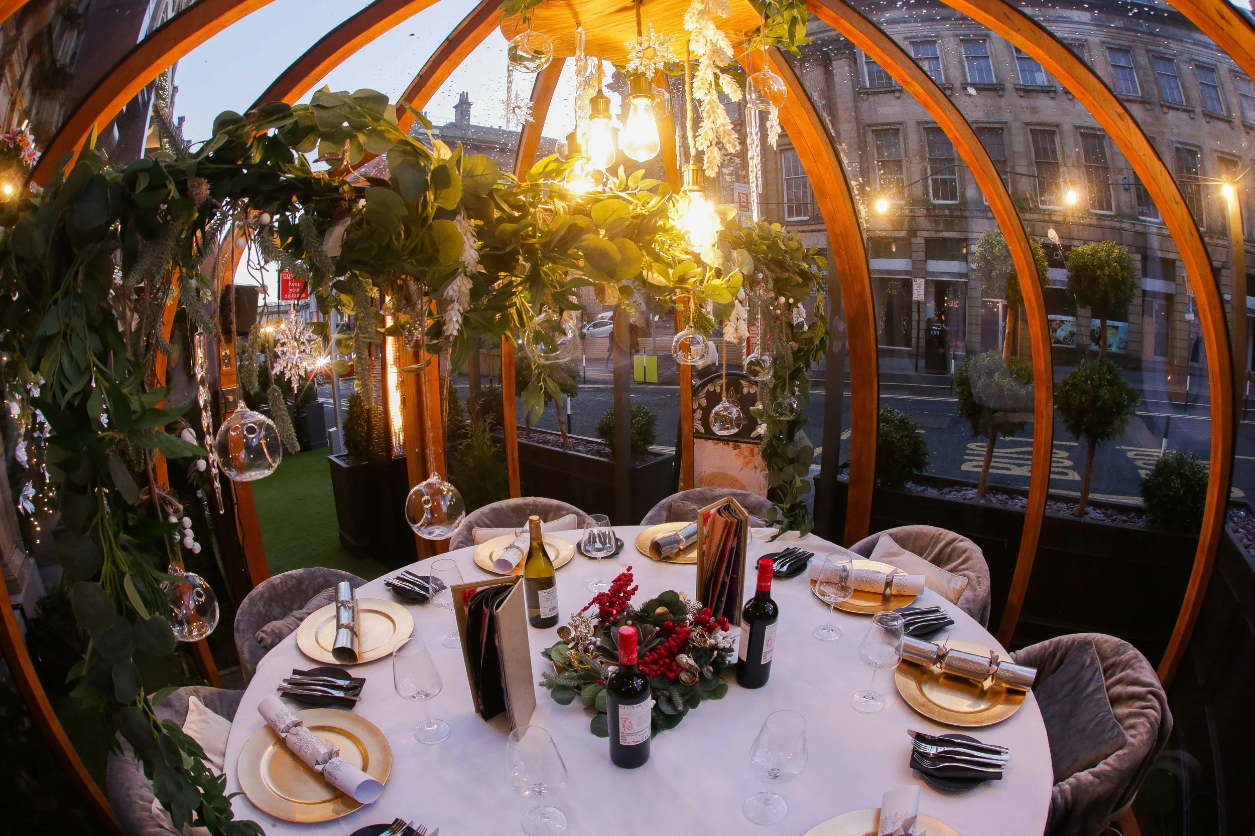 A round dining table set for a celebration inside a glass-greenhouse with wooden framing. The table has gold-rimmed plates, wine bottles, wine glasses, and napkins. Decorative greenery, red berries, and Christmas ornaments serve as the centerpiece. Hanging glass orbs and warm light bulbs create a festive ambiance, with city buildings visible outside.