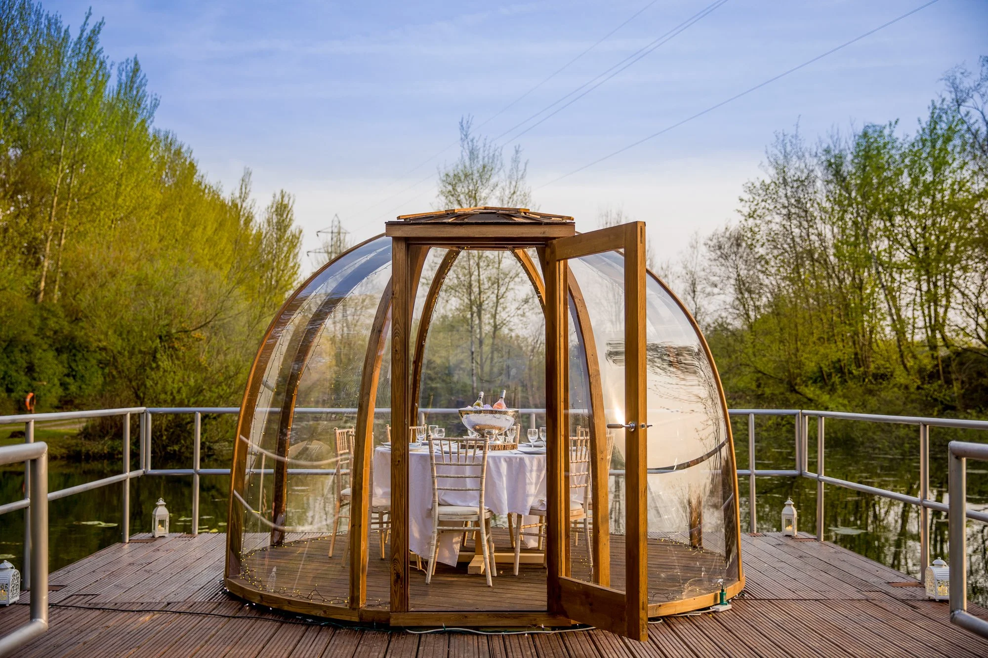 A wooden and glass gazebo with a door open, set on a wooden deck overlooking a body of water and surrounded by trees, with a table set for dining inside.
