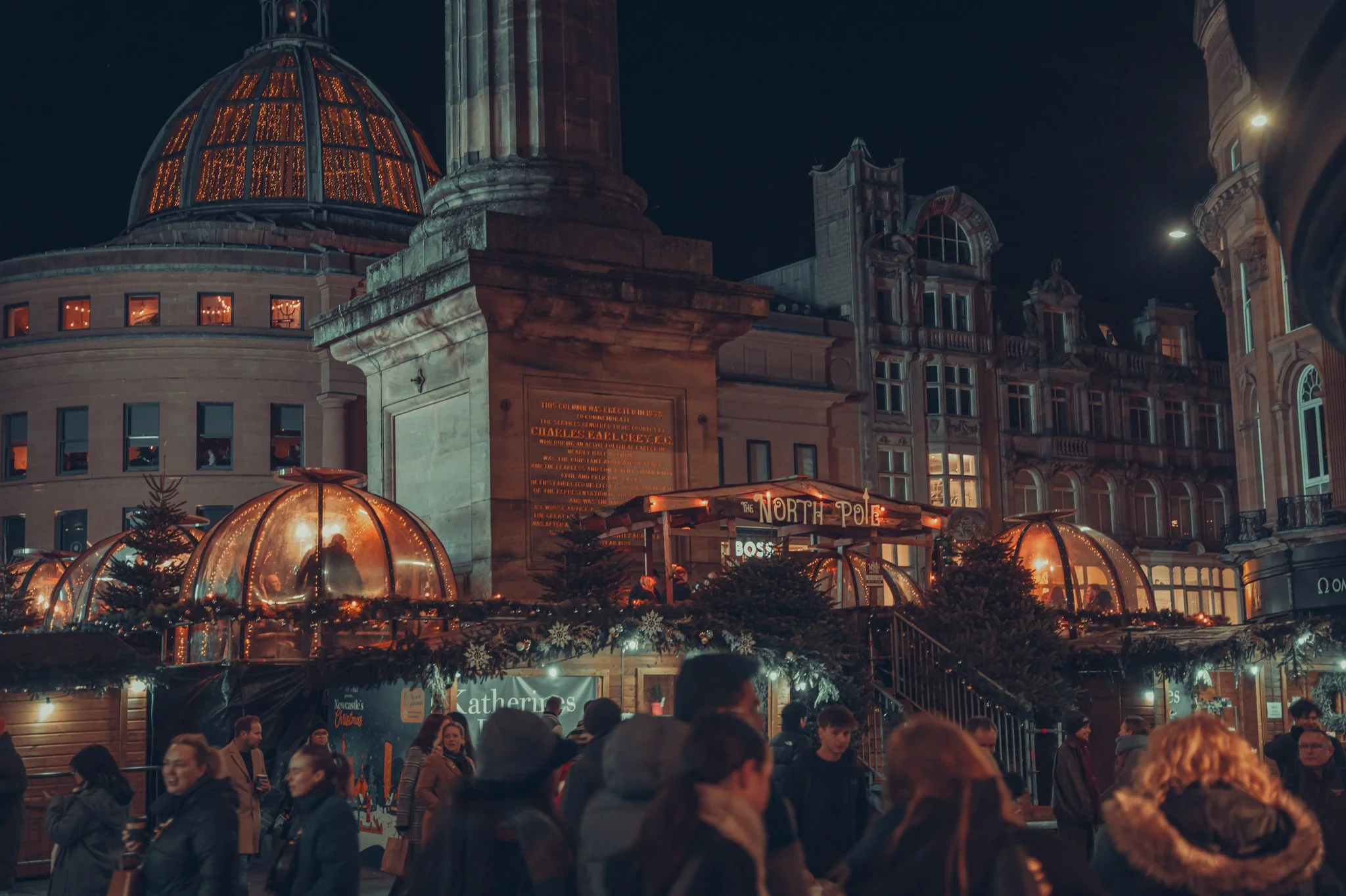 Nighttime scene at a Christmas market with people gathered in front of a large historic building with a domed roof, decorated with lights and festive decorations, including trees and illuminated signs.