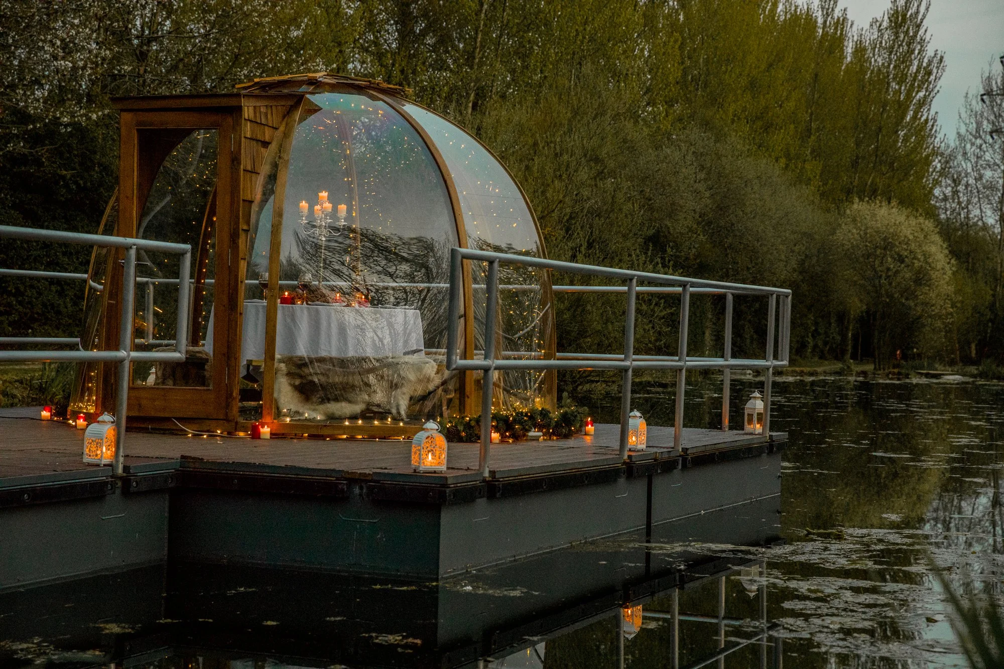 A floating glass-enclosed dining area on a dock by the water, decorated with candles and string lights, with a table set and a chandelier inside, surrounded by trees.