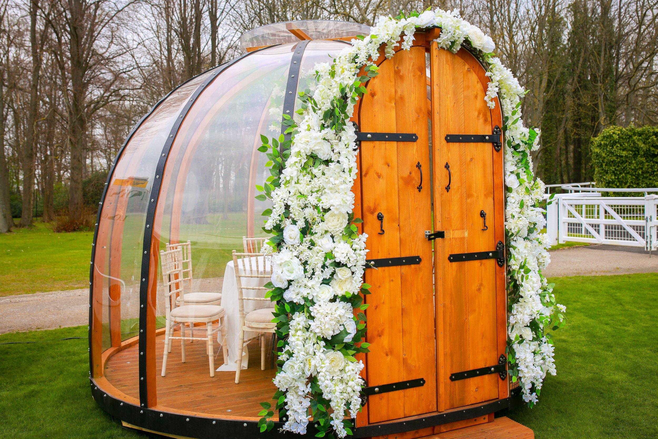 A small wooden greenhouse decorated with white flowers, set up outdoors with tables and chairs inside.