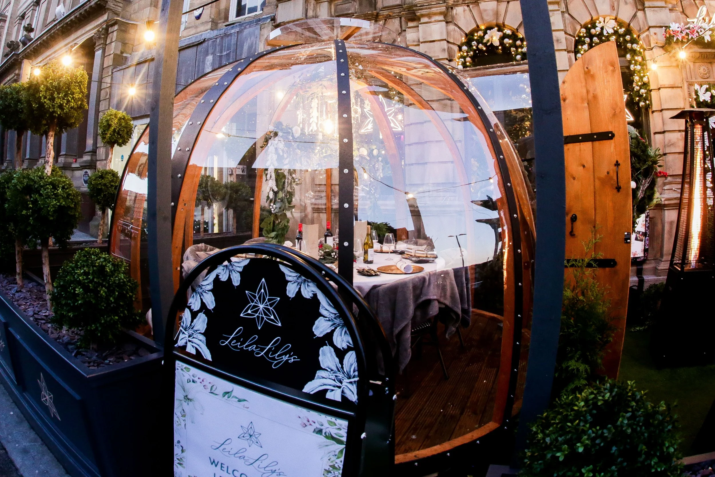 A cozy outdoor dining space enclosed in a transparent, geodesic dome with string lights, decorated with potted plants and Christmas garlands, set up on a city street for a festive dinner, with a sign reading Leila Lily's.