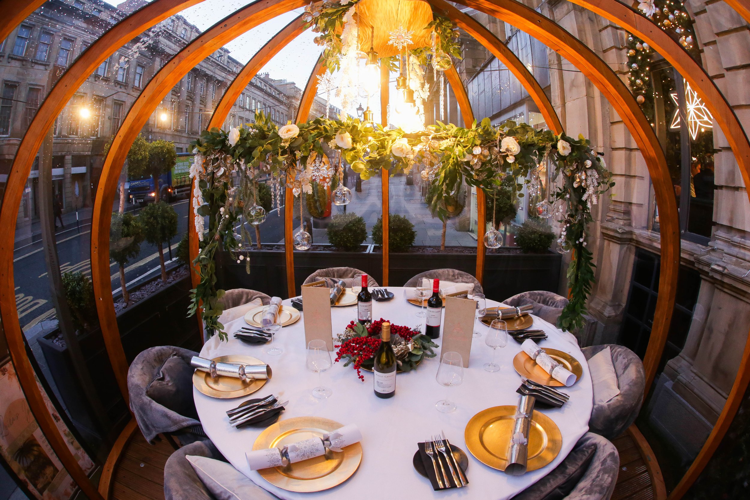 Round dining table set for a holiday celebration inside a glass-enclosed structure with wooden arches, decorated with Christmas ornaments, greenery, candles, wine bottles, and tableware.