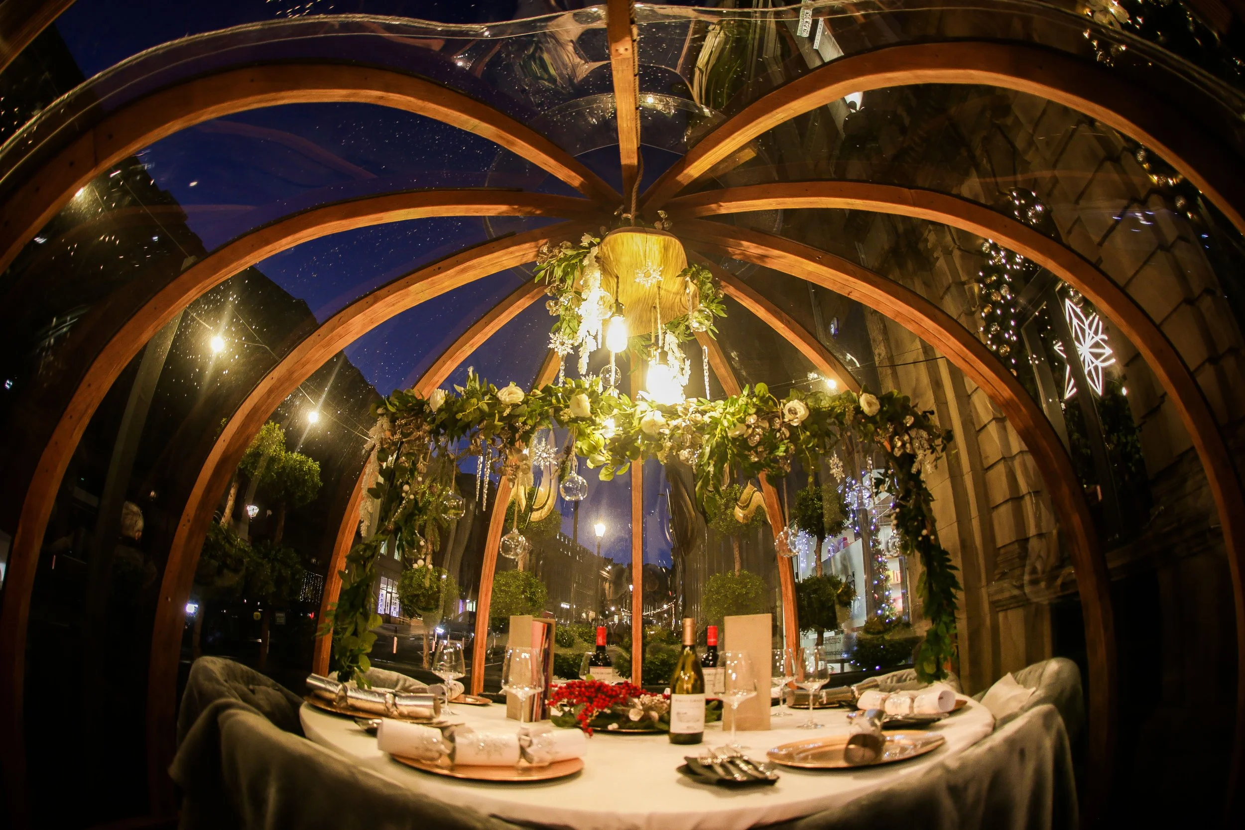 Inside a glass-domed, decorated for a festive dinner with a round table set with wine bottles, glasses, wrapped gifts, and a floral centerpiece, under a wooden arch with hanging lights, with holiday snowflake decorations visible outside.