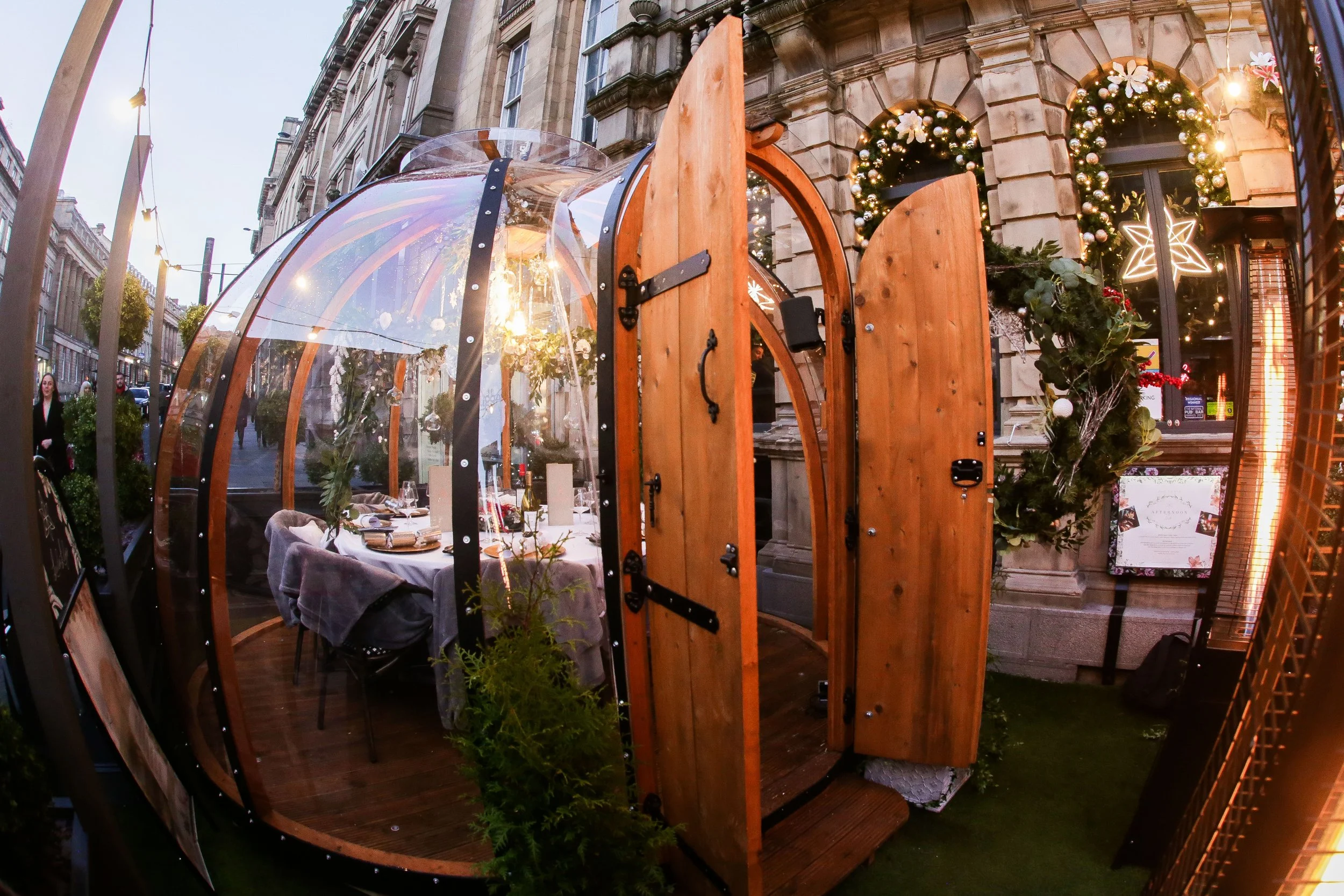 A glass igloo dining setup on a city street, decorated for Christmas with wreaths, lights, and star-shaped ornaments.