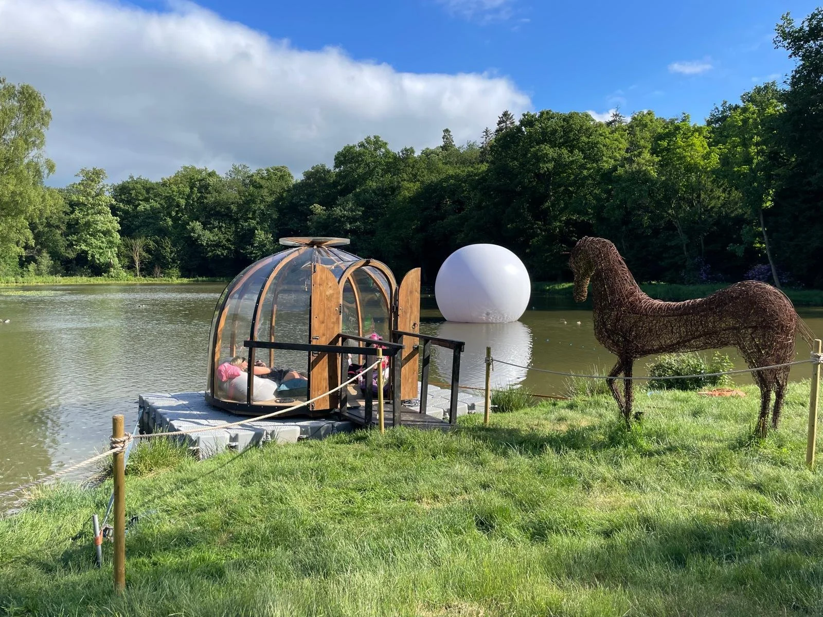 A small glass and wood structure with a person lying inside on a raft, float near a lakeside. There is a large white sphere and a wireframe horse sculpture on the grassy shore, with trees and a partly cloudy sky in the background.