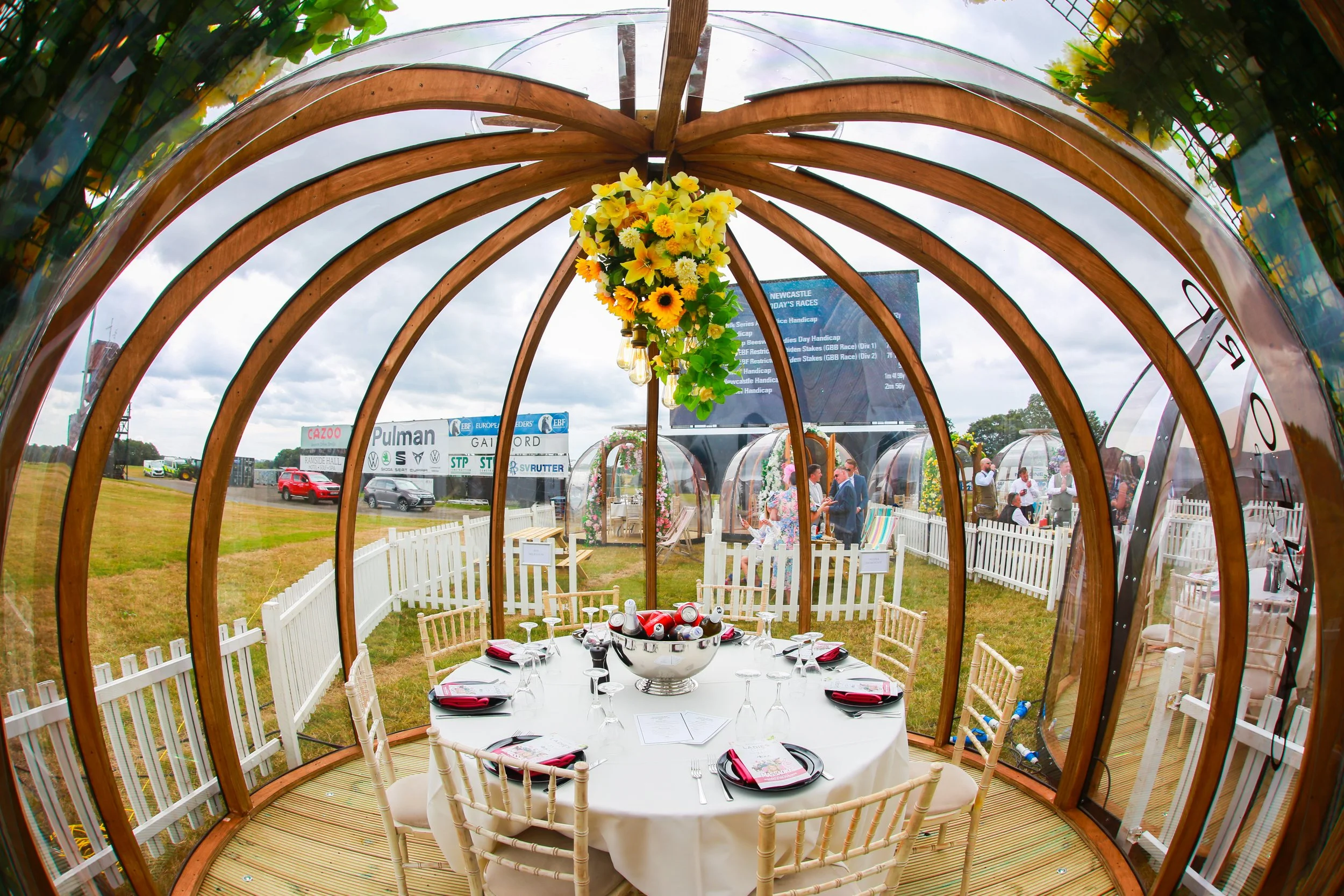 Round table set with black plates, glasses, and red napkins inside a glass and wood dome structure at an outdoor event, with yellow flowers hanging from the top center of the dome.
