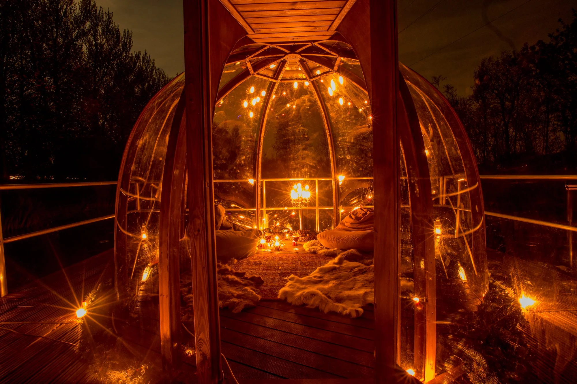 A cozy, illuminated glass and wood pod on a deck at night, decorated with string lights and plush rugs.