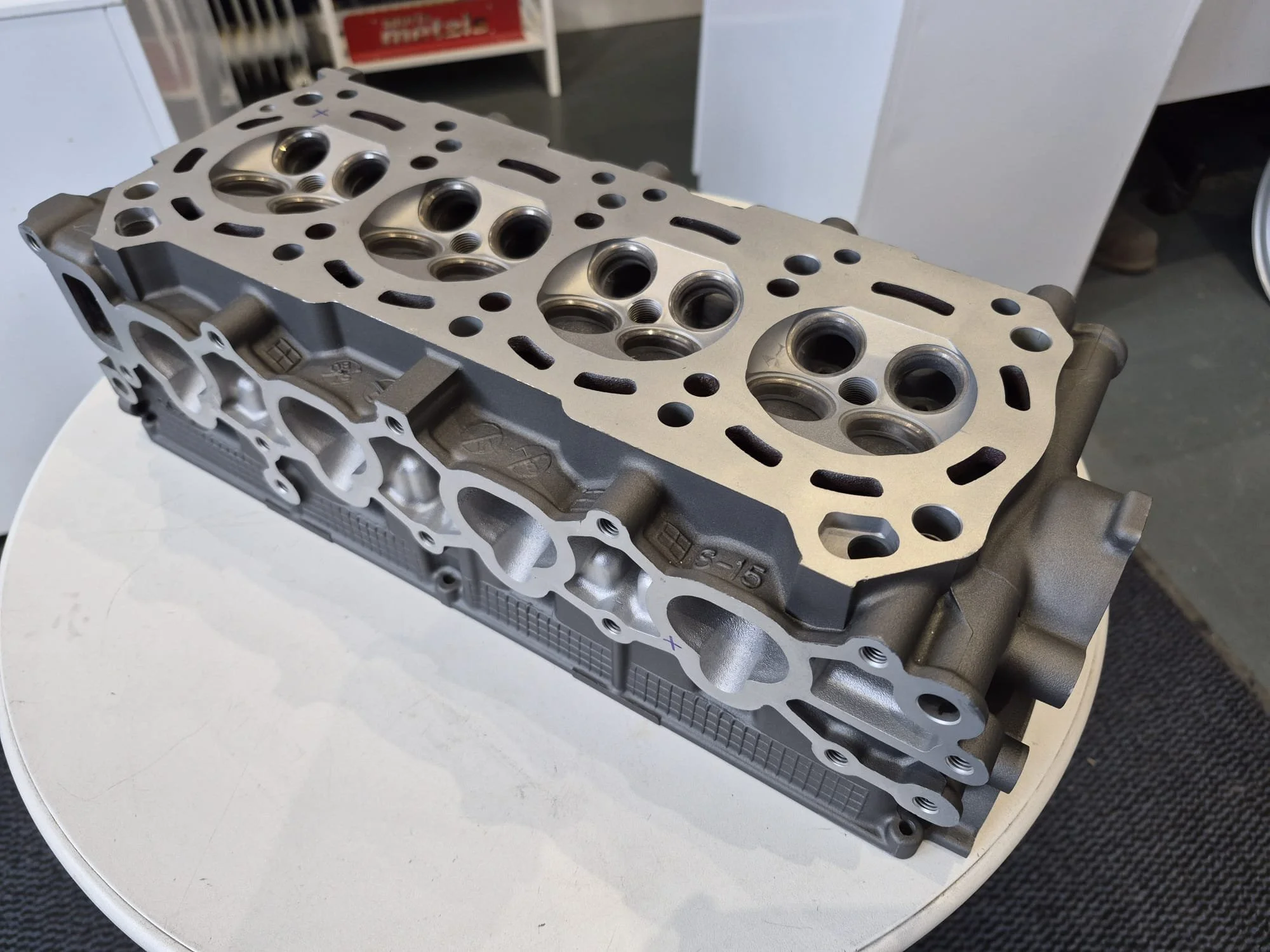 A new metal engine cylinder head with multiple intake and exhaust ports on a white circular table in a workshop setting.