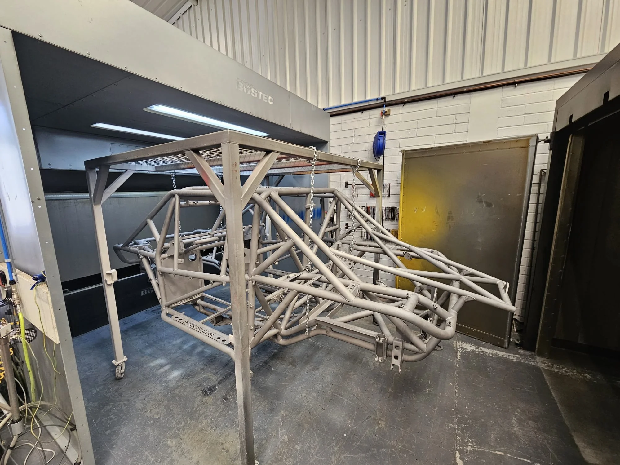 Metal chassis frame on a wheeled stand inside a workshop, with a large black industrial machine in the background.