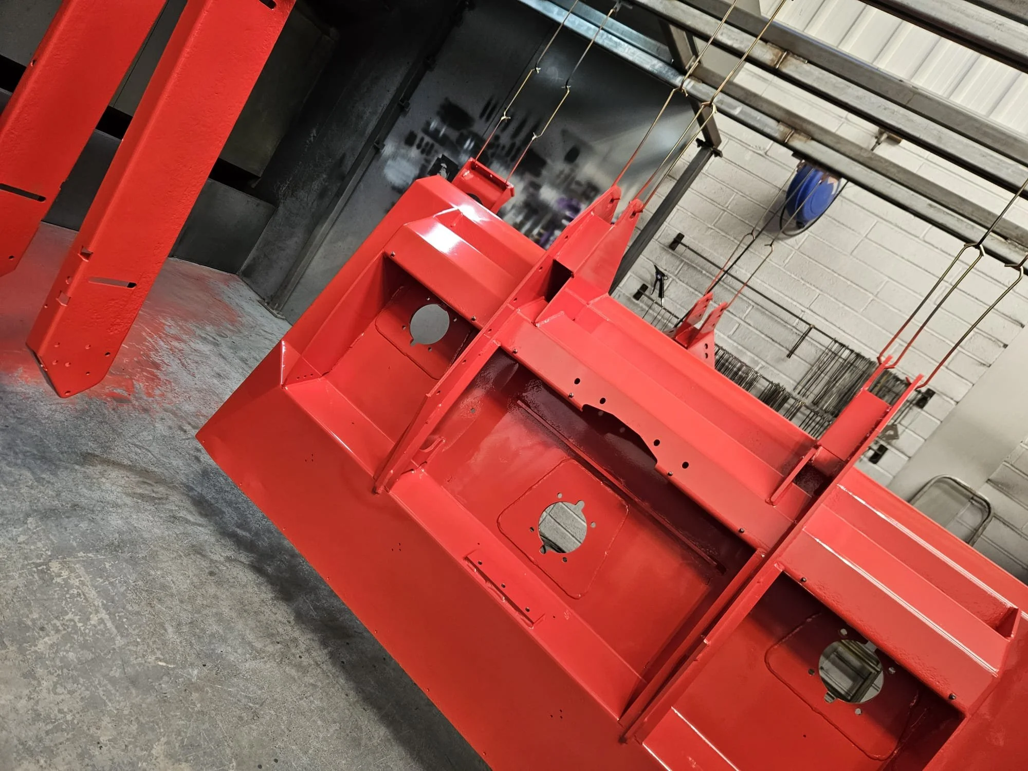 Red-powdercoated metal structure, possibly a vehicle part, hanging in a workshop or industrial setting with tools and equipment visible in the background.