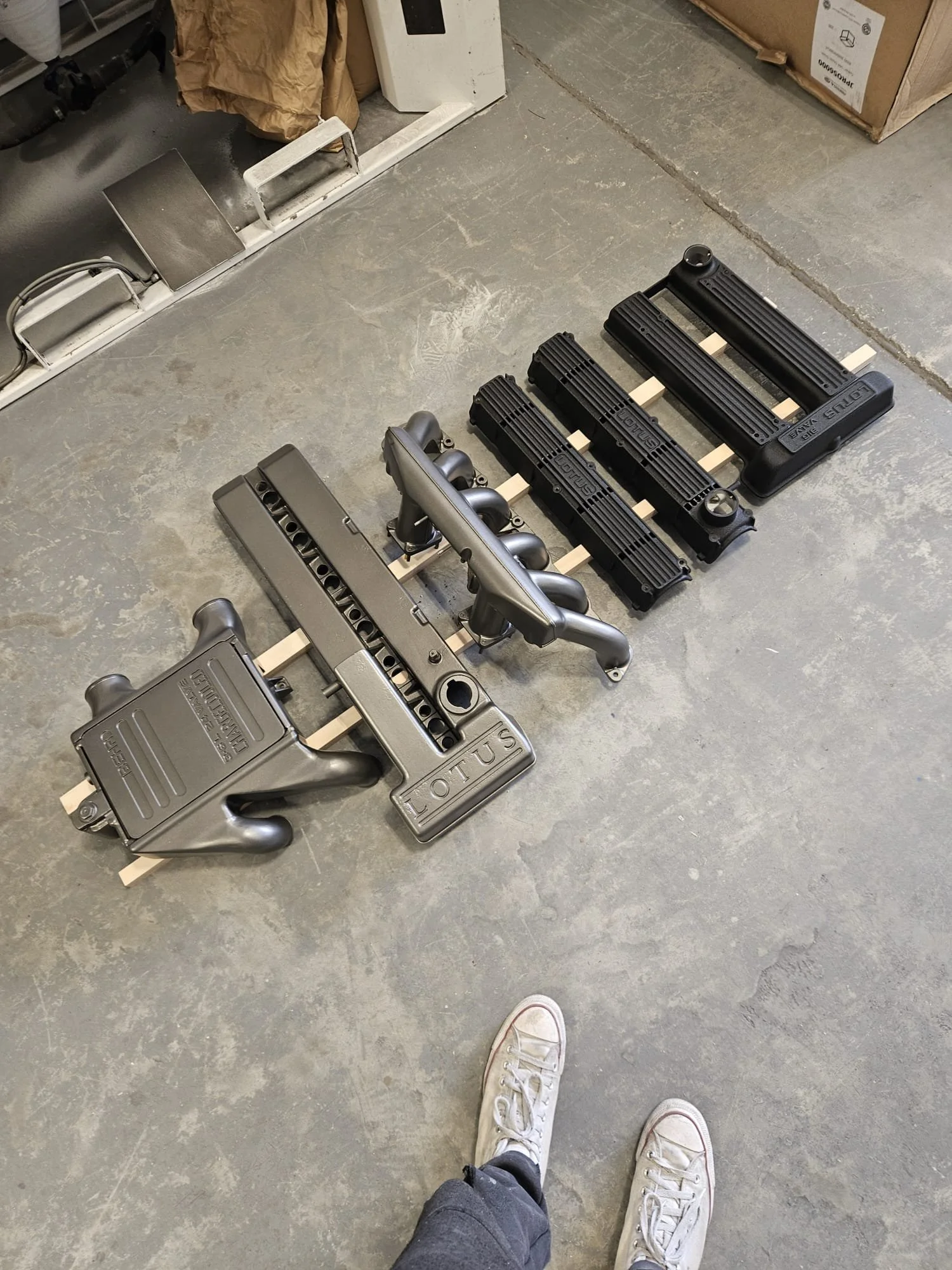 Various engine parts, including intake manifolds, valve covers, and intake plenum, laid out on a concrete floor in a workshop.