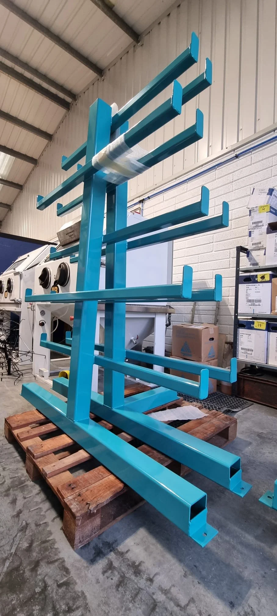 Blue powder coated metal rack with multiple horizontal shelves, placed on a wooden pallet in an industrial warehouse setting.