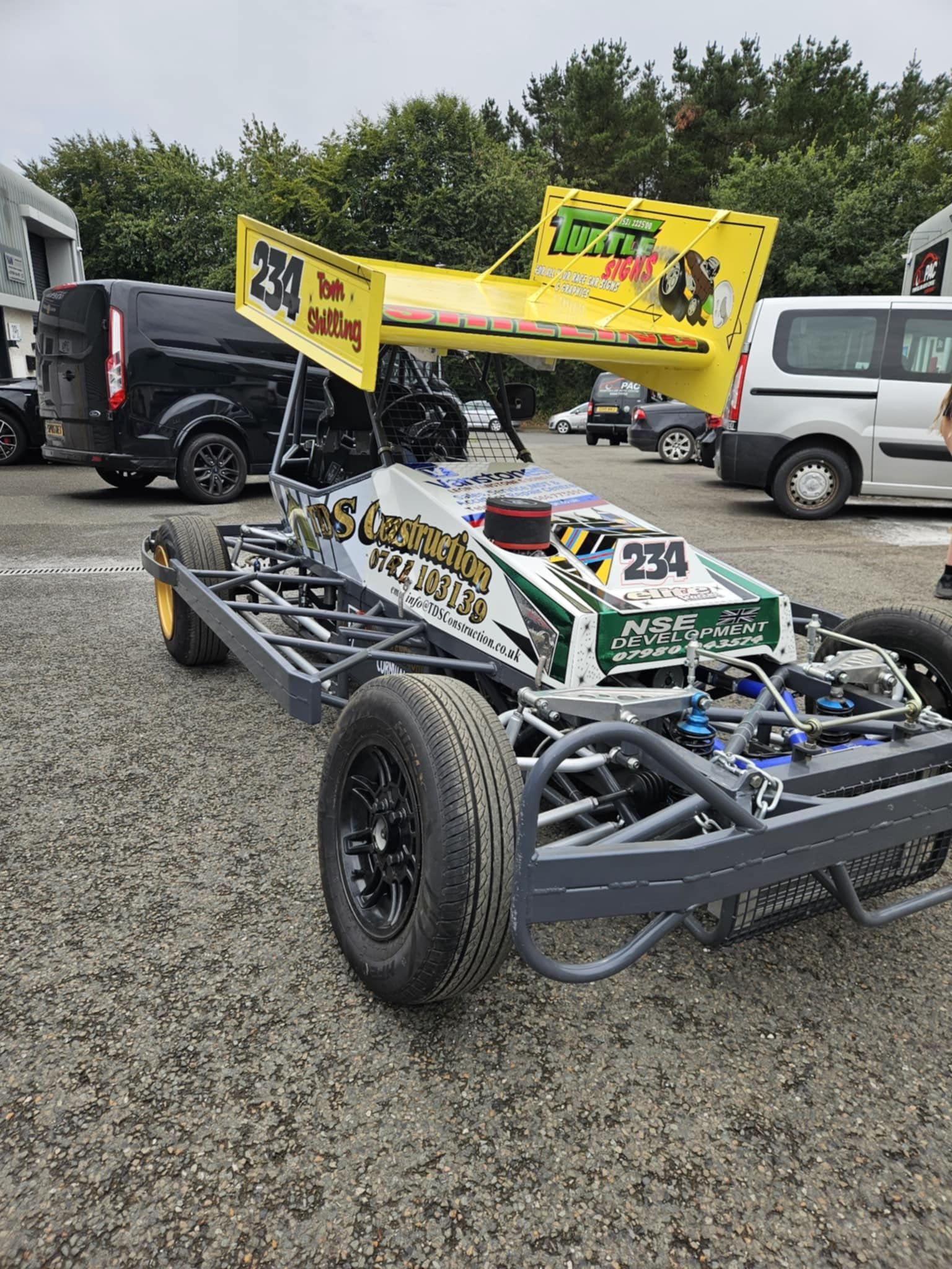 A go-kart with the number 234, featuring a yellow rear wing with tournament signs, parked on a gravel lot with other vehicles in the background.