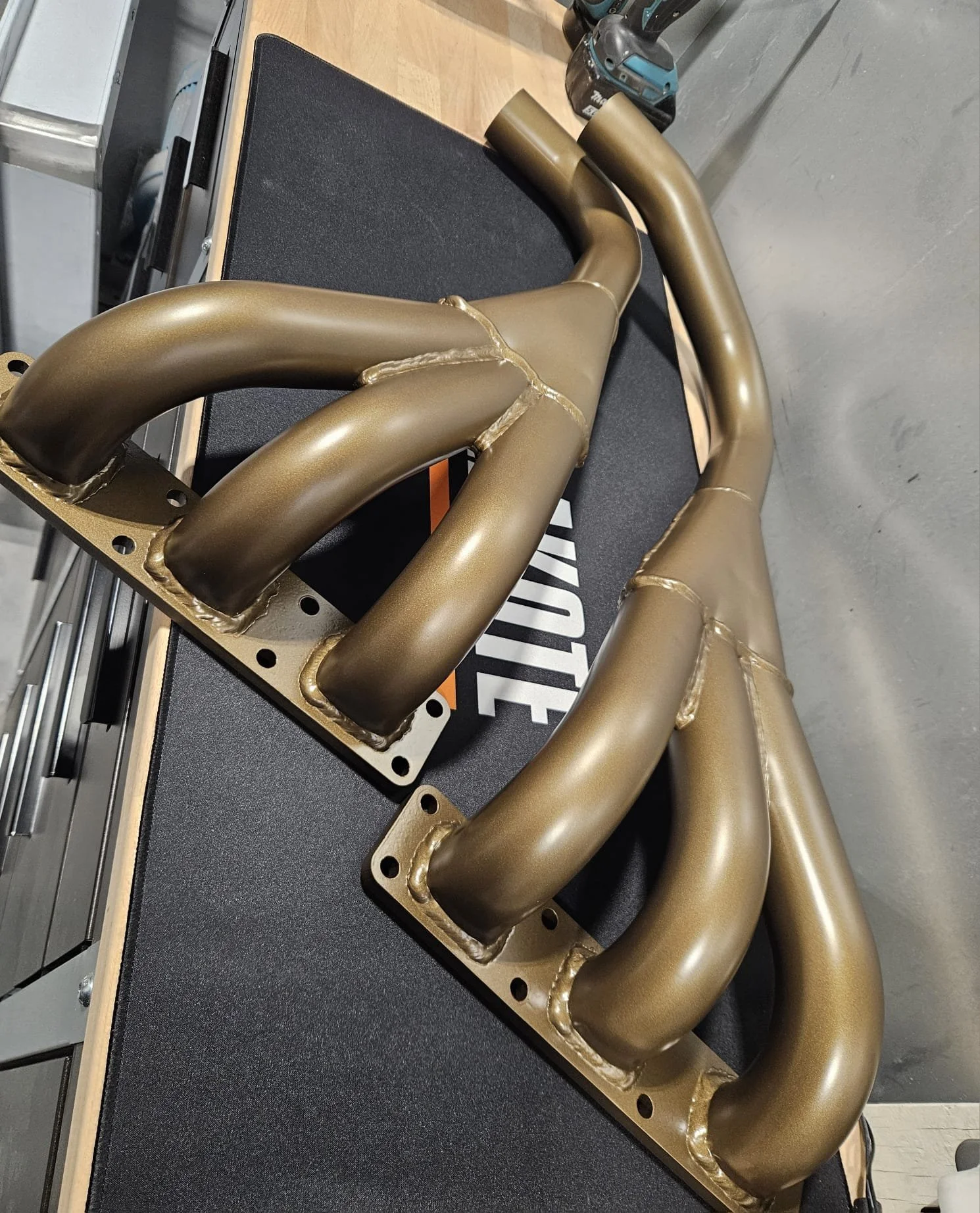 A metallic automotive exhaust header with multiple curved pipes, mounted on a black surface showing craftsmanship and painting expertise. 