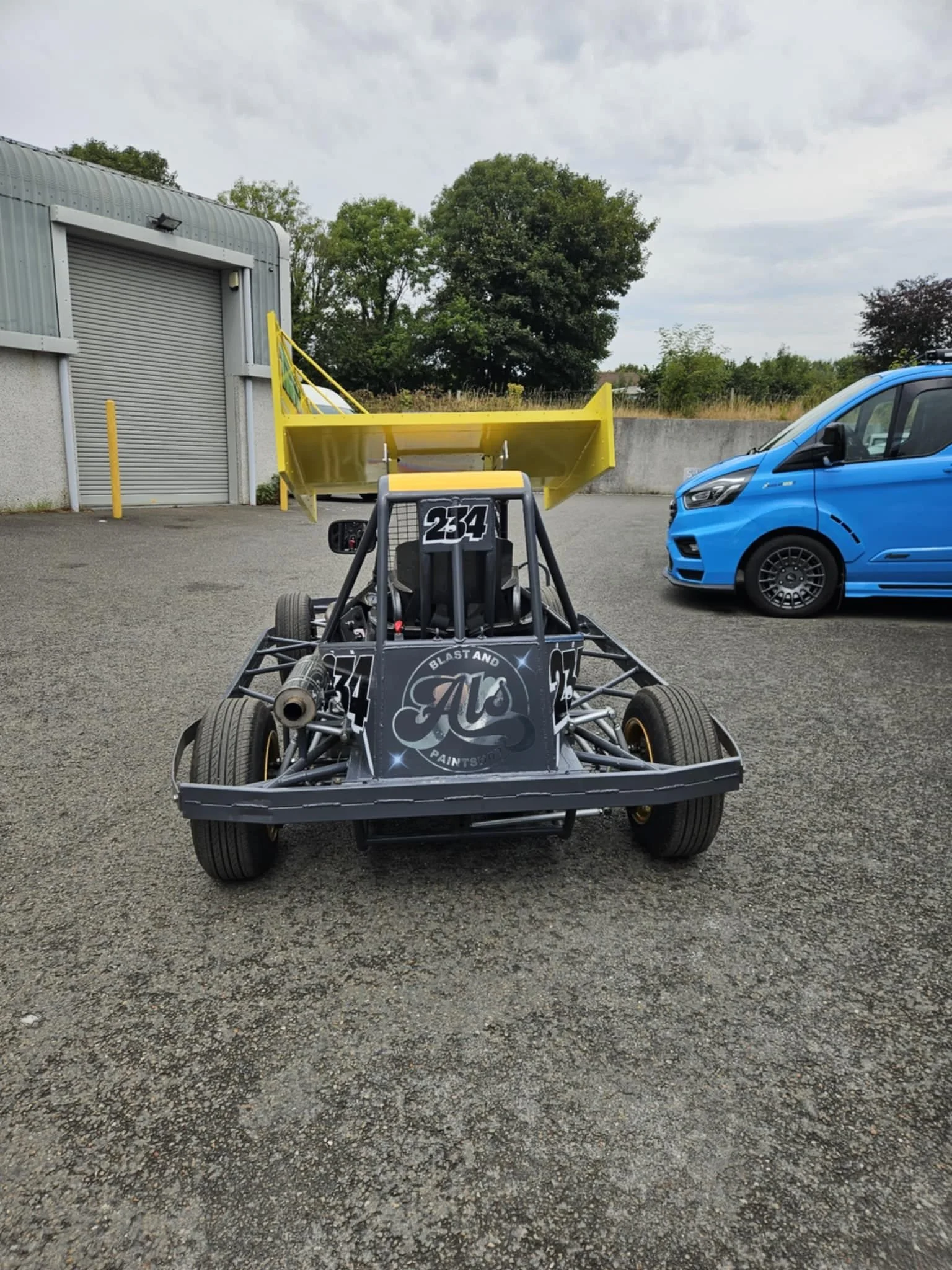 A racing go-kart with the number 234 on the front, parked on an asphalt lot, with a yellow wing and a sponsor logo on the front panel, next to a blue van.