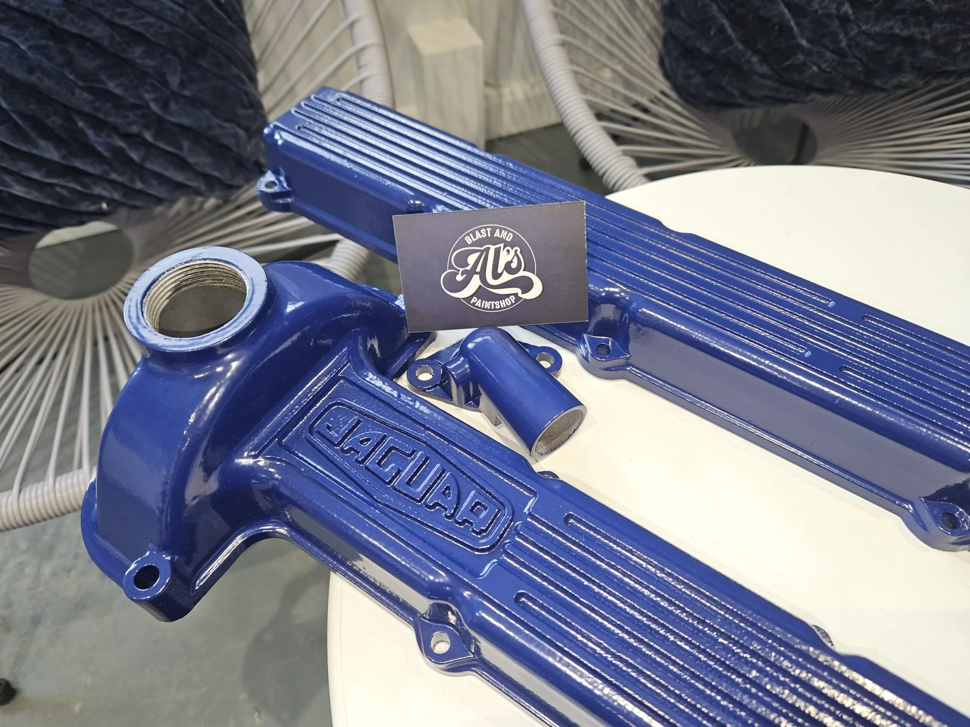Blue automotive parts, including an intake manifold and valve cover, are placed on a white surface with a black business card from Al's Blast and Paintshop positioned on top. The background contains chairs with circular woven designs.