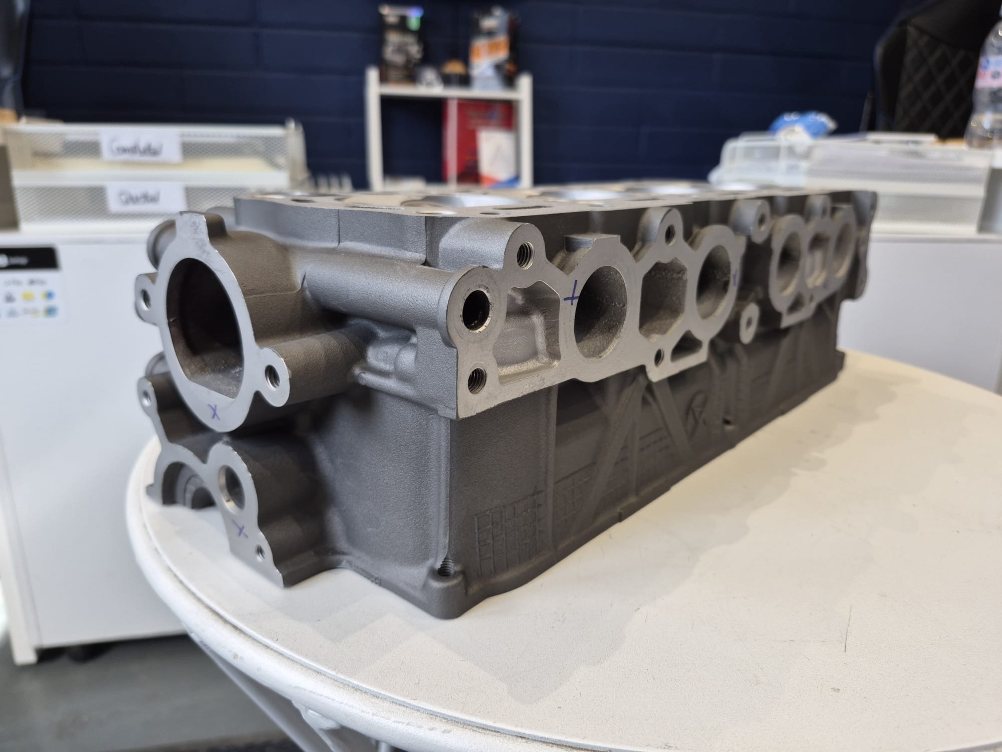 A metallic engine cylinder head placed on a white table in an indoor setting with shelves and bottled water in the background.