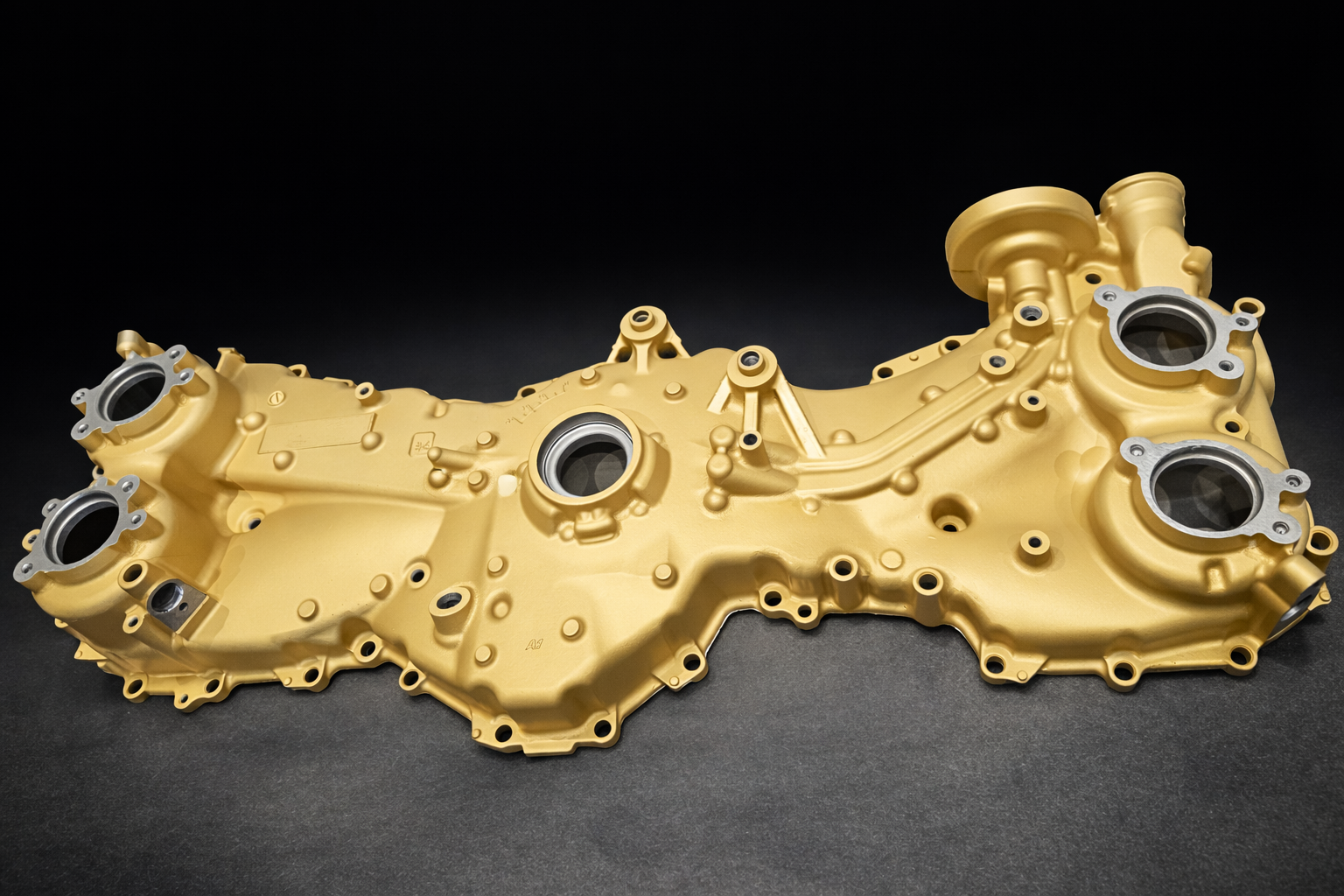 Golden-colored engine cover with multiple circular openings and bolt holes, placed on a dark surface against a black background.