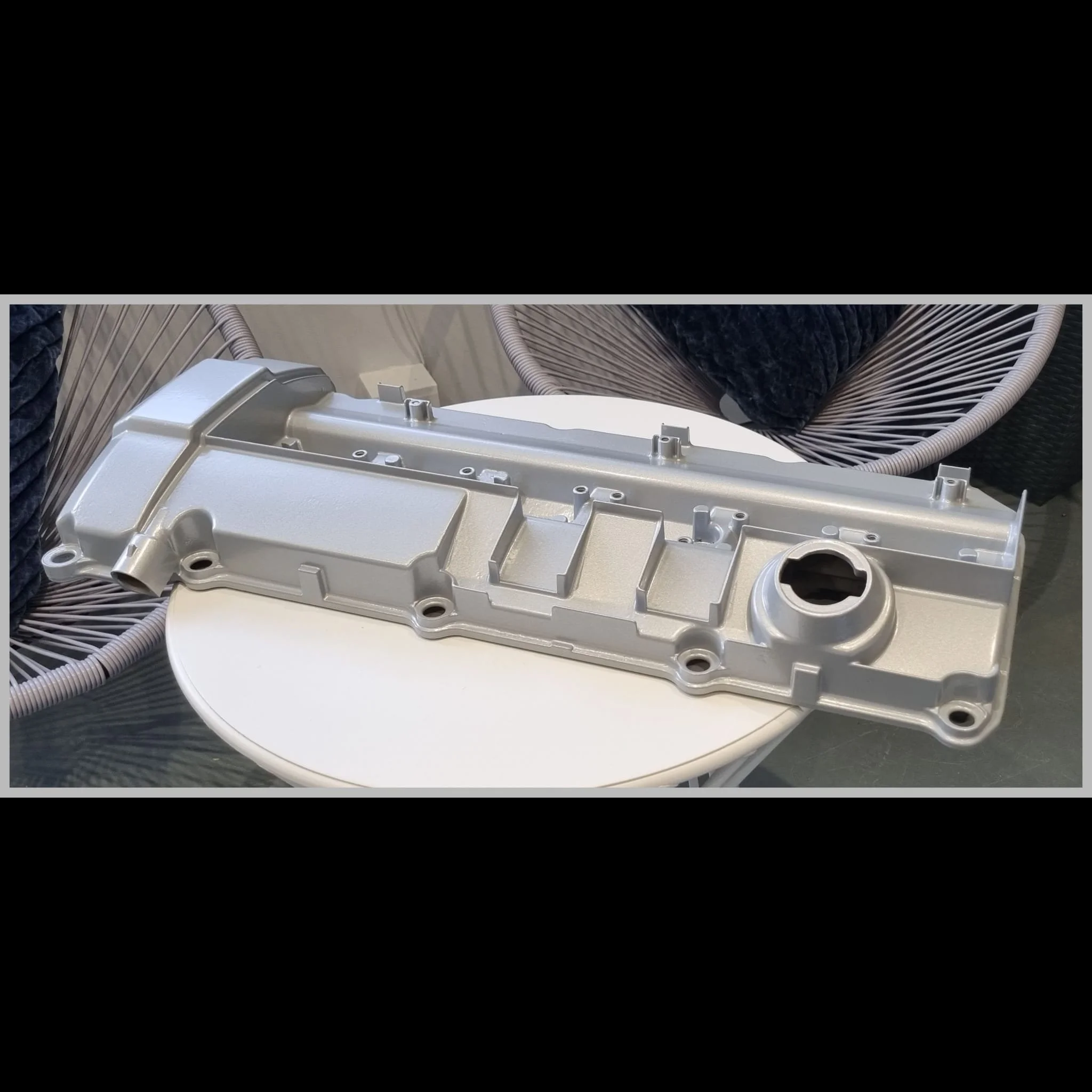 Aluminum engine valve cover on a white circular table, with woven chairs and cushions in the background.