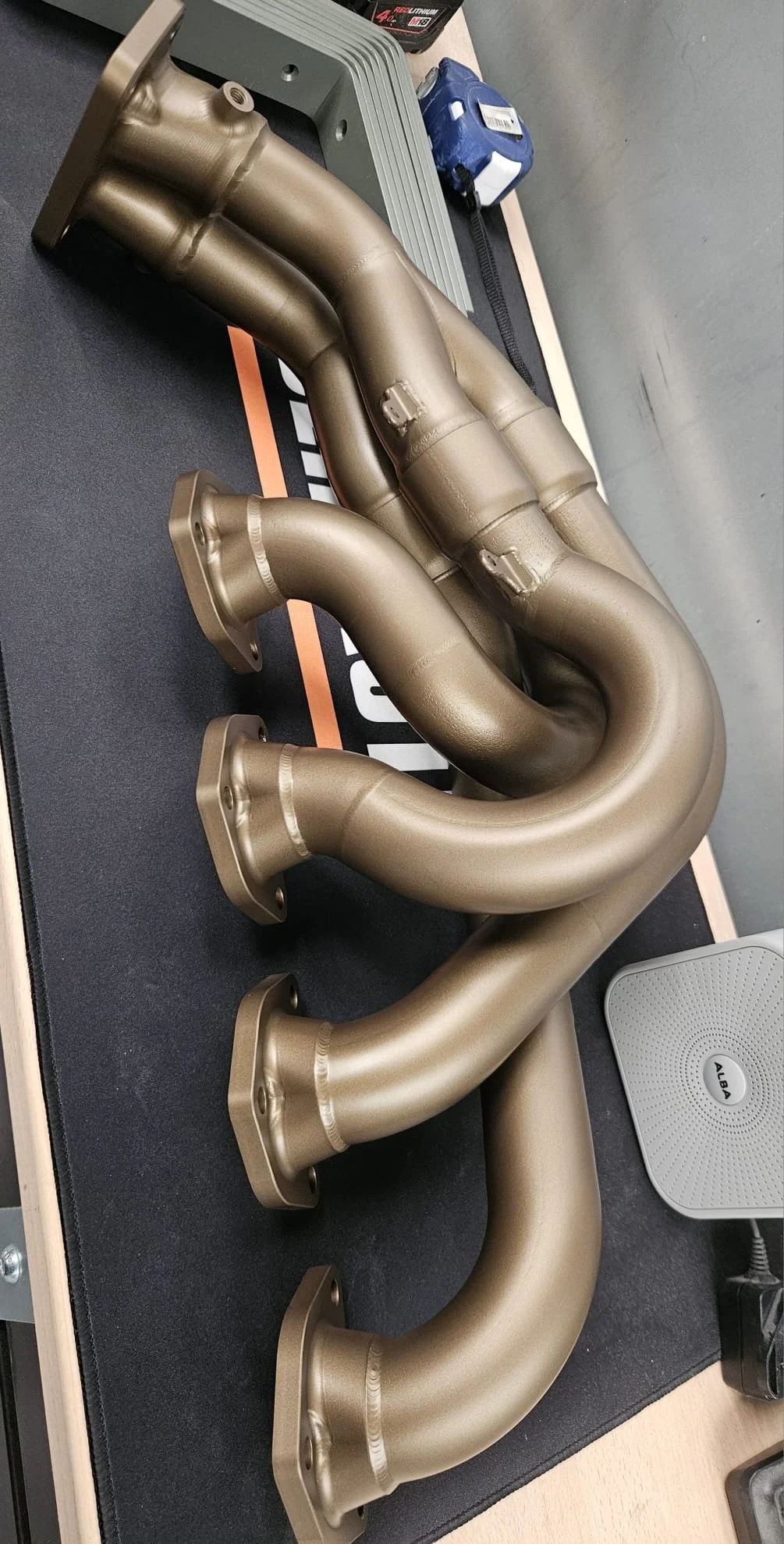 A metallic car exhaust header with multiple pipes, placed on a black workspace mat finished in a gold finish - possibly cerakote