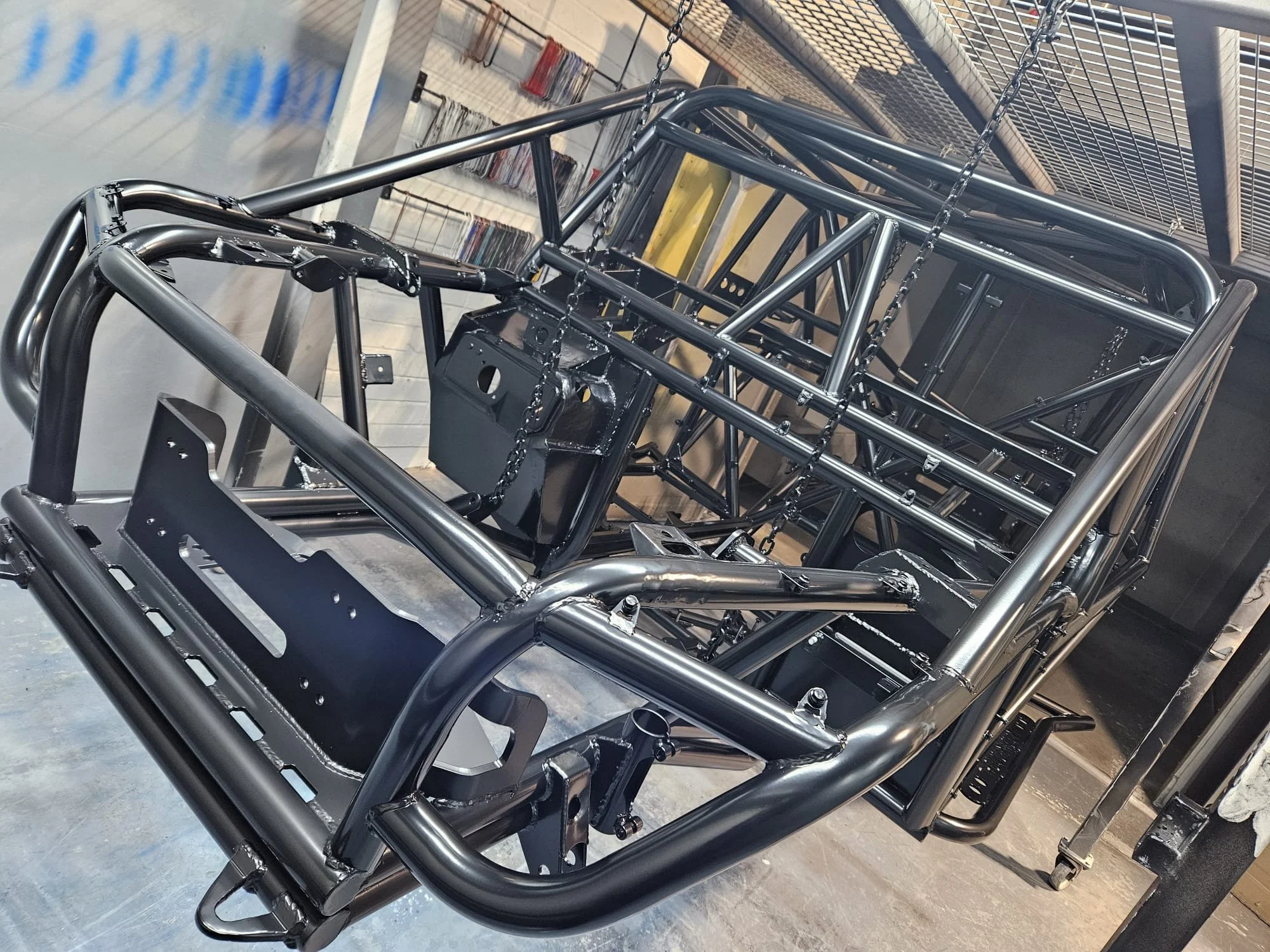 A metal frame of a race car or off-road vehicle in a workshop, with a black dashboard and wiring visible.