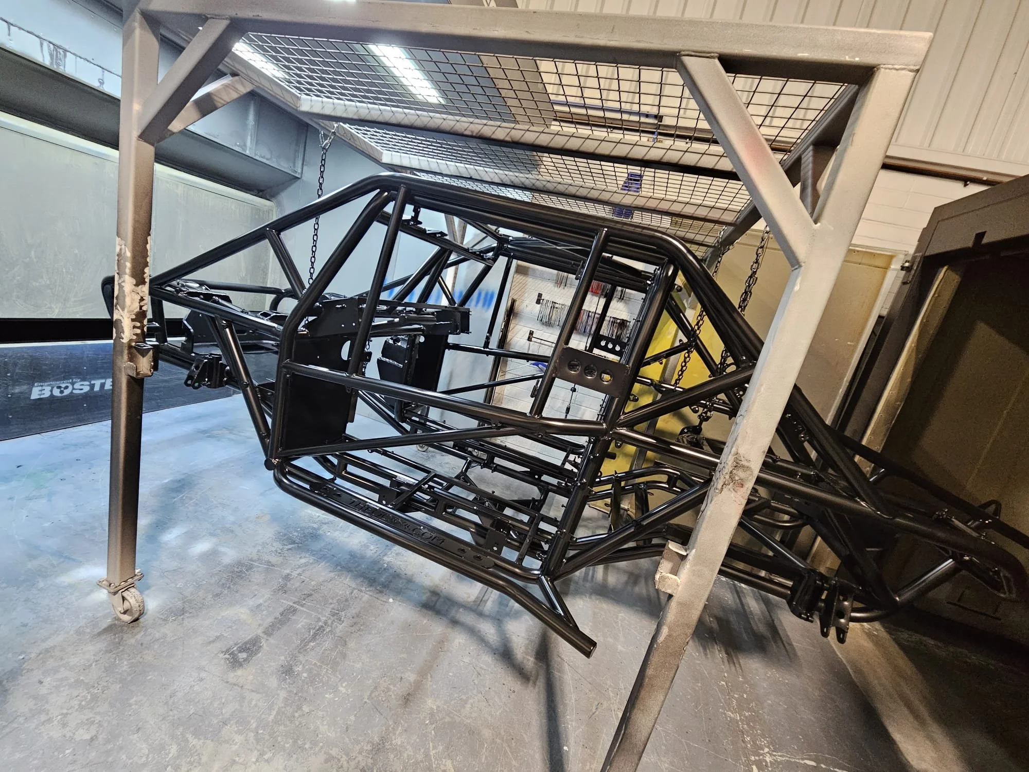 A metal frame roll cage for a race car is positioned on a wheeled metal stand in a workshop or garage.