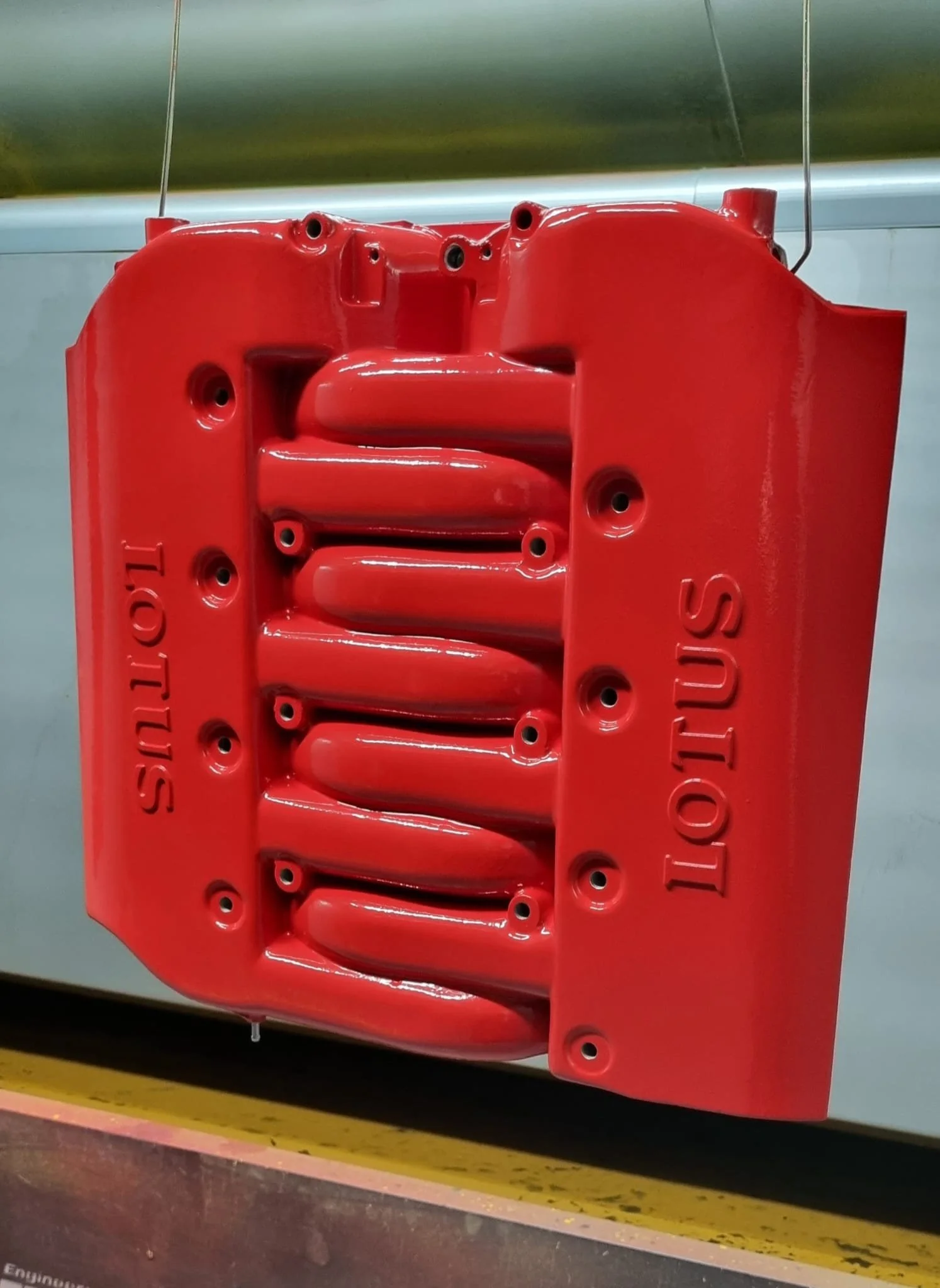 Red engine valve cover with the words 'lotus' on one side and 'lotus' on the other, hanging inside a factory or workshop.