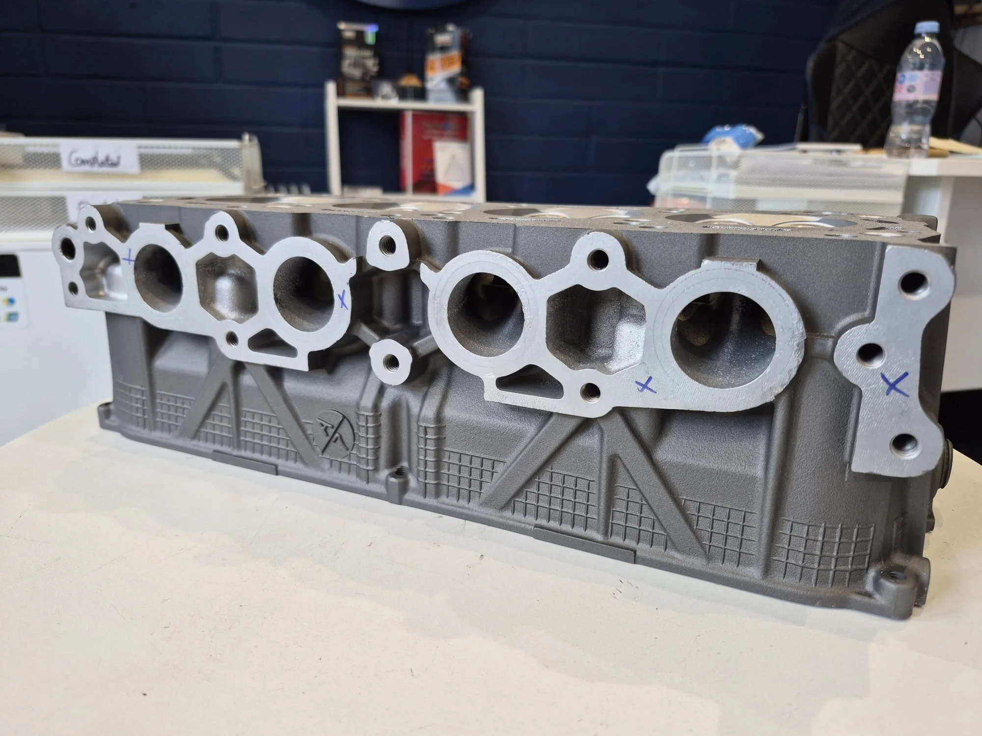 A gray engine cylinder head with machined holes and markings, placed on a white surface in a workshop or manufacturing setting.