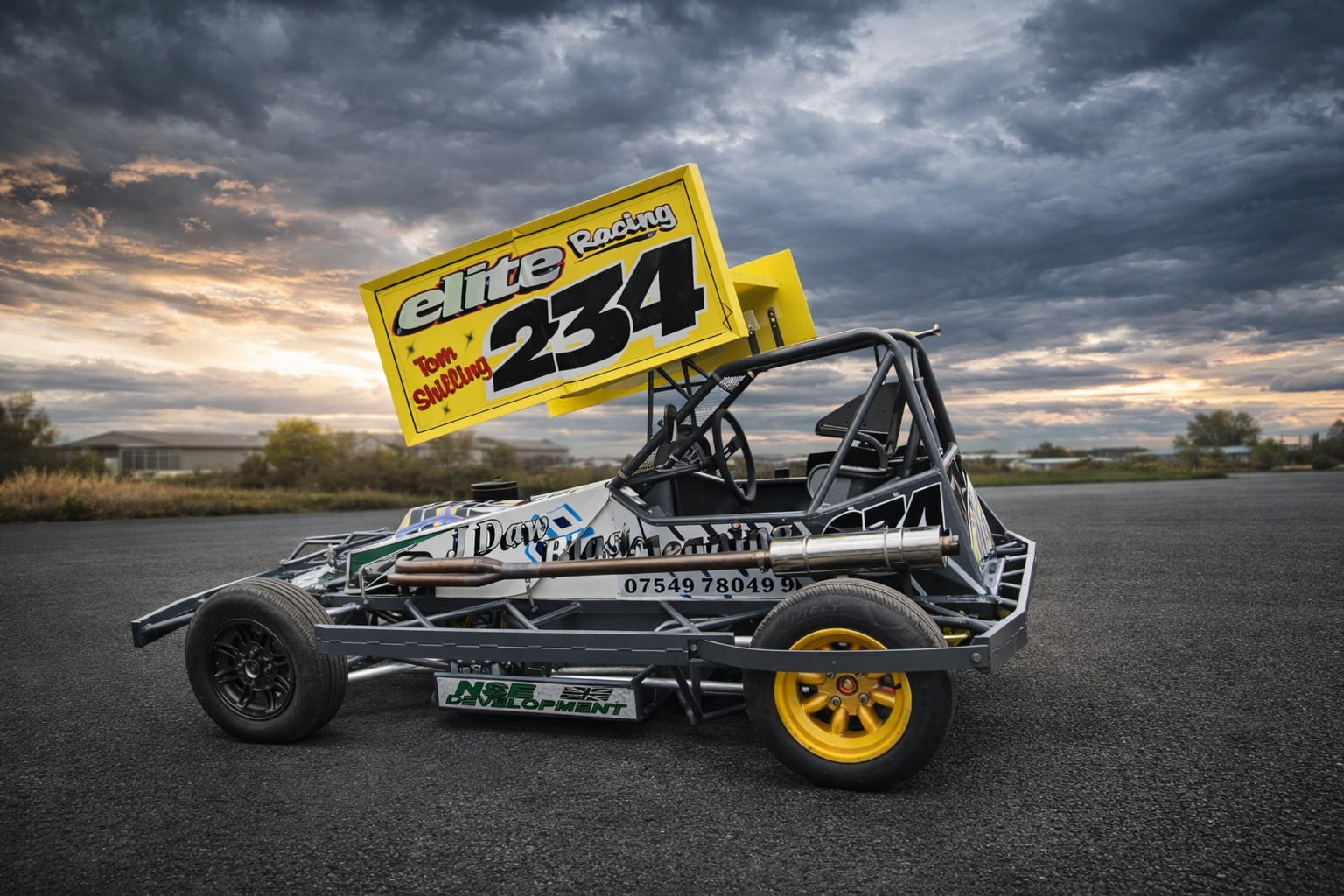 a small race car with a yellow wing, black tires, and a metallic frame, parked on an asphalt surface against a cloudy sky during sunset.