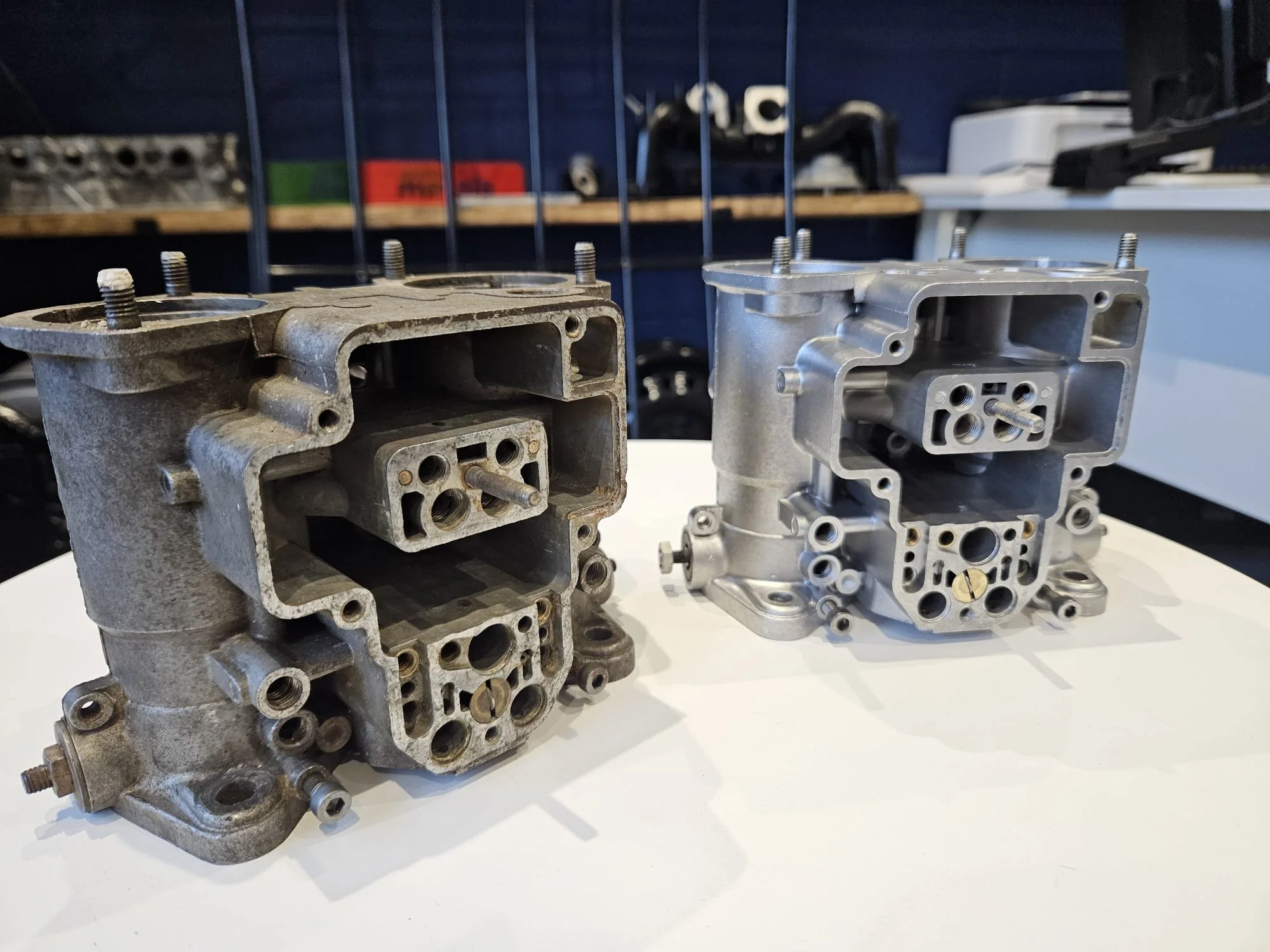 Two metal engine cylinder heads placed on a white surface with a workshop background showing restoration work with a before and after showing the vapor work.