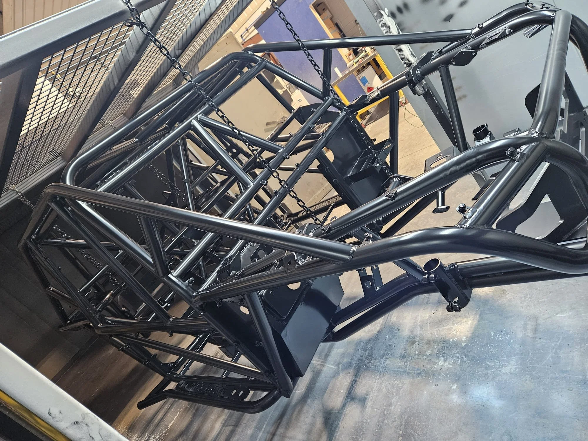 Metal frame structure of a race car or similar vehicle, with various metal tubes and chains, in a workshop or manufacturing setting.