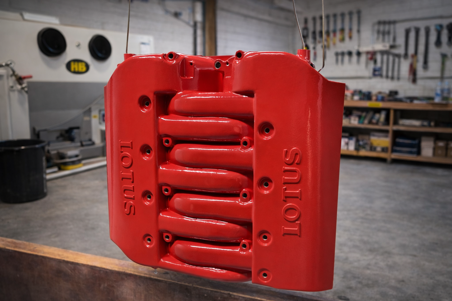 A red metal engine component with the words 'LOTUS' molded into it, in a workshop with tools and shelves in the background.