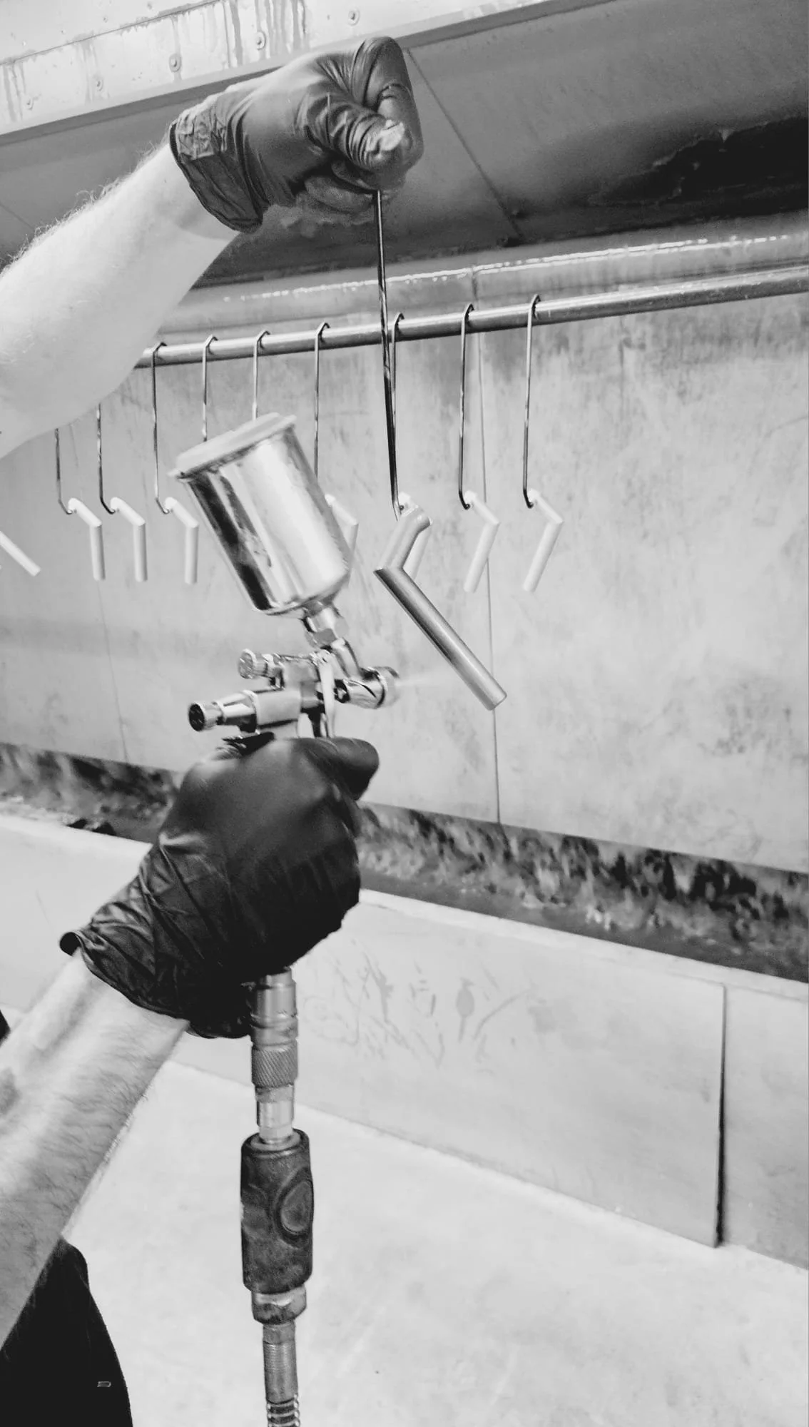 A person wearing black gloves spray painting metal hooks hanging on a rail in a workshop or industrial setting.