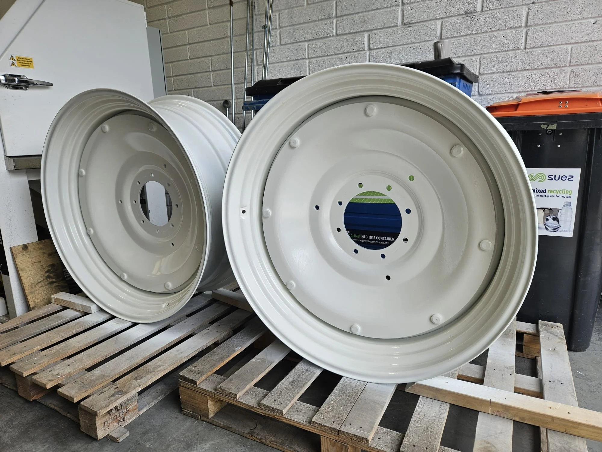 Two white painted / powder coated metal tractor wheels resting on wooden pallets in an industrial setting.