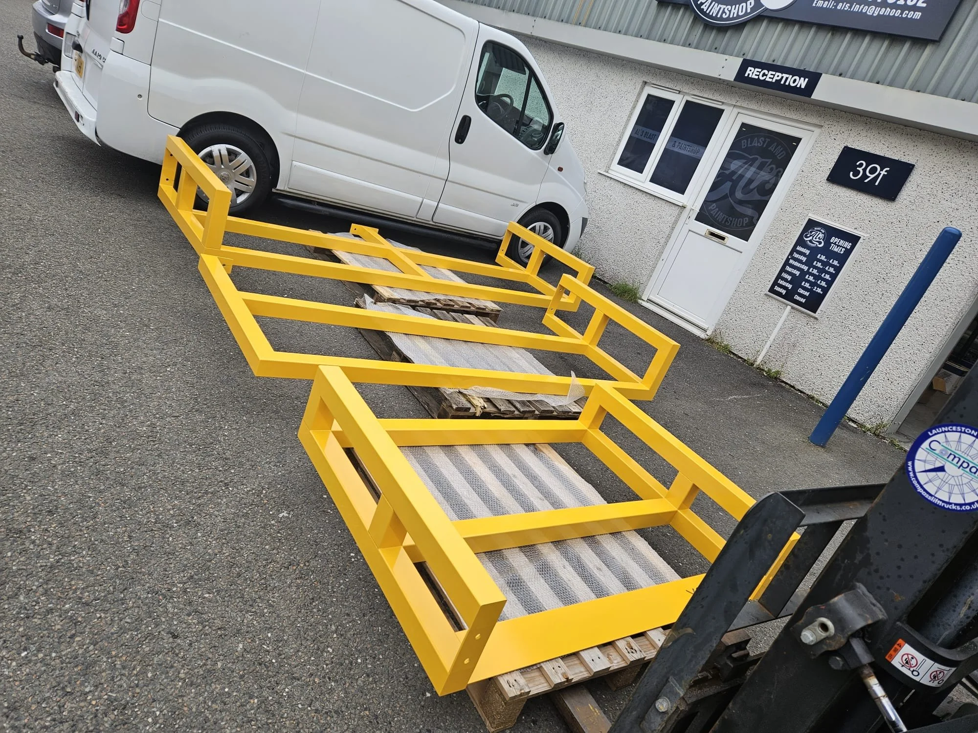 A white van parked beside a yellow metal frame on a pallet outside a paint shop with signs and a building in the background.