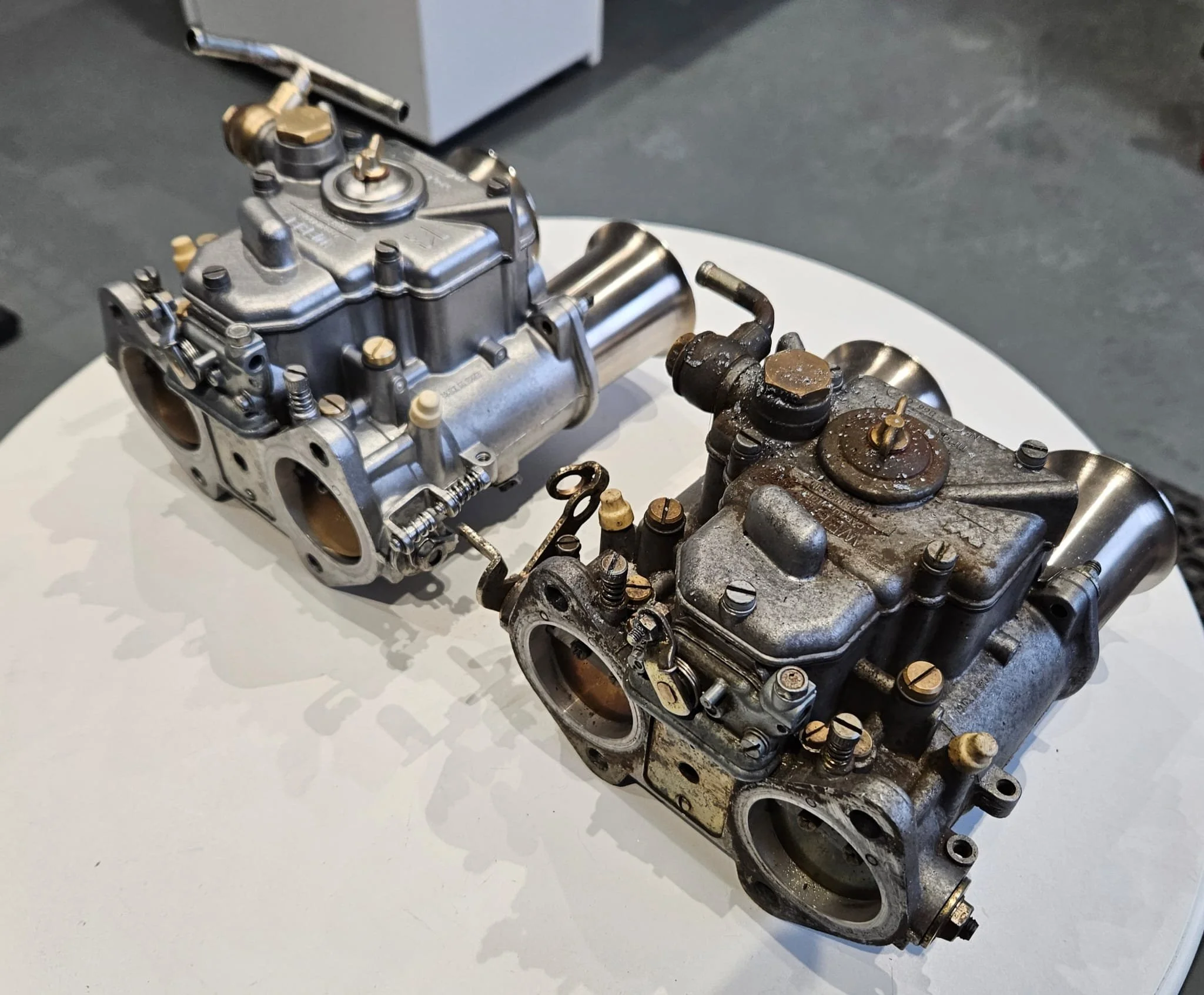 Comparison of two vintage carburetors, one clean and shiny, the other rusty and dirty, placed on a white surface showing a before and after of vapour blasting.