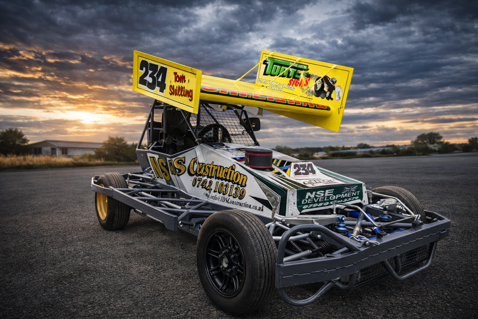 Race car with a large yellow wing and black tires parked on an empty asphalt surface at sunset, with clouds in the sky and buildings visible in the background.